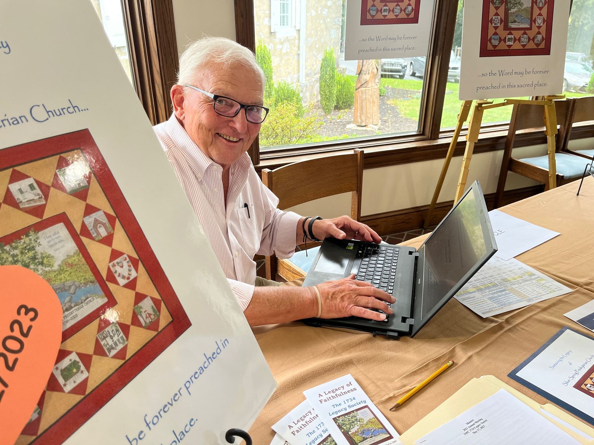 A man sitting at a table using a laptop with a sign behind him that says 2023