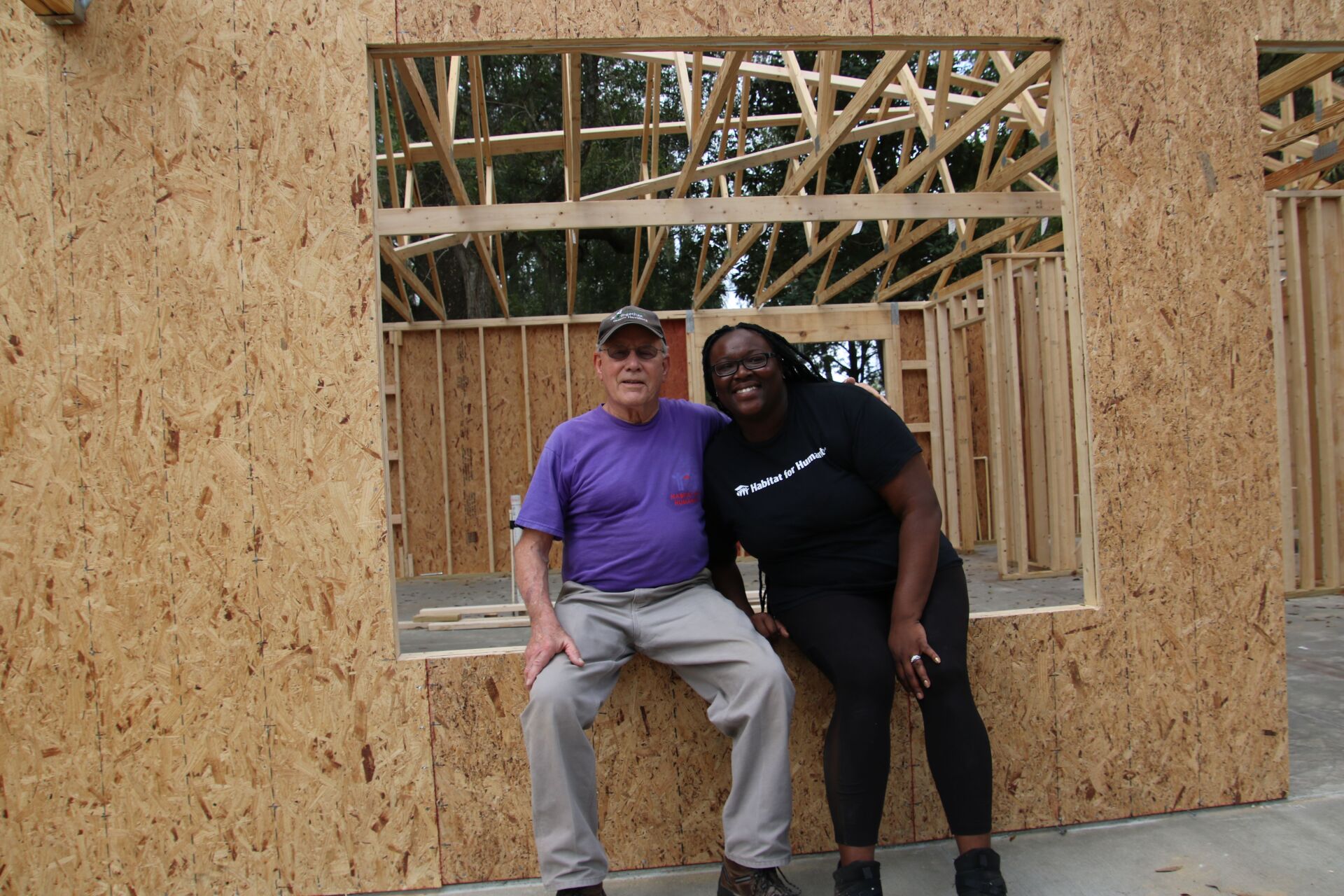Two people smiling, posing in front of the wooden frame of a building under construction.