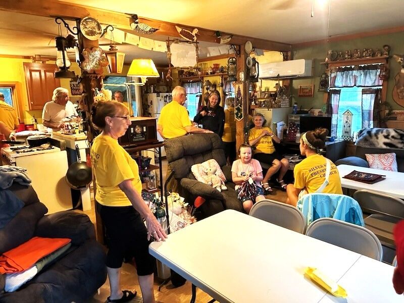 People in yellow shirts gather in a cluttered room with tables. Some hold cards and laugh.