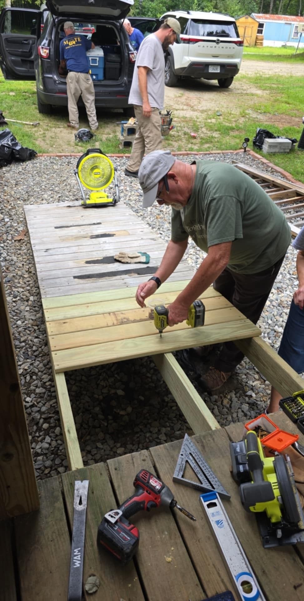 Men building a wooden structure outdoors. One man uses a drill. Tools and cars are visible.