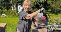 Woman outdoors with safety glasses, holding a circular saw and smiling.