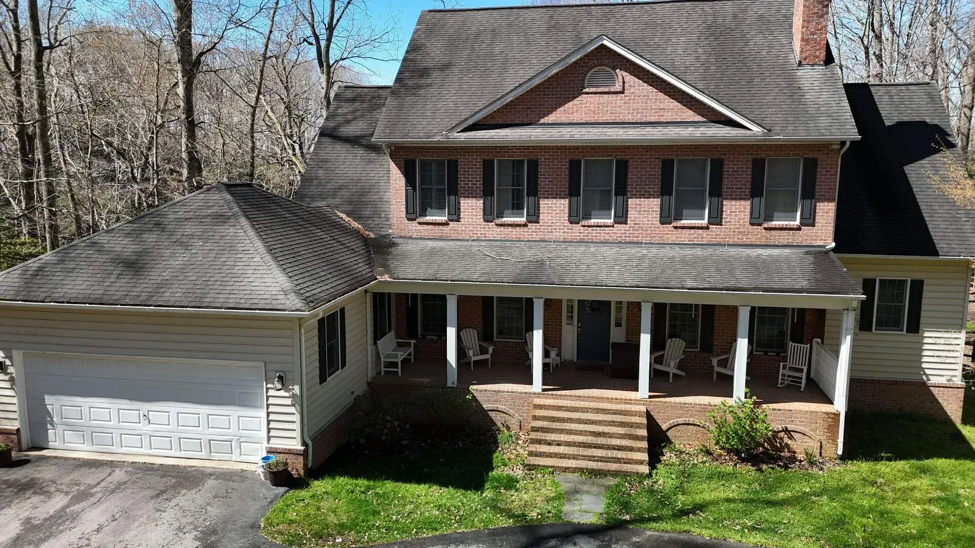 Two-story brick house with white garage, front porch, and dormer windows surrounded by trees