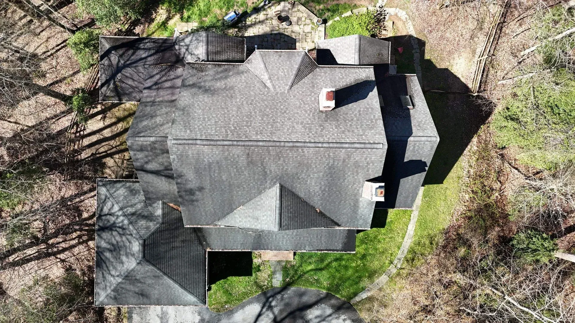 Aerial view of a dark-roofed house surrounded by trees, with a small chimney and green yard.
