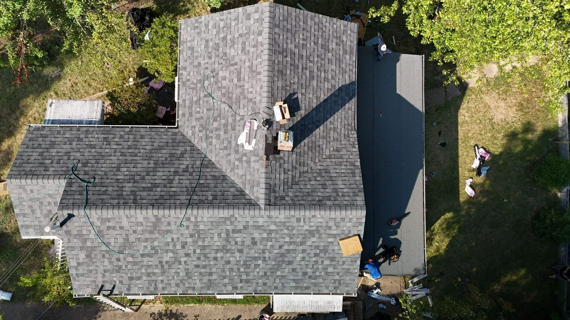 Aerial view of a gray-shingled house roof with workers and tools near the chimney and side yard