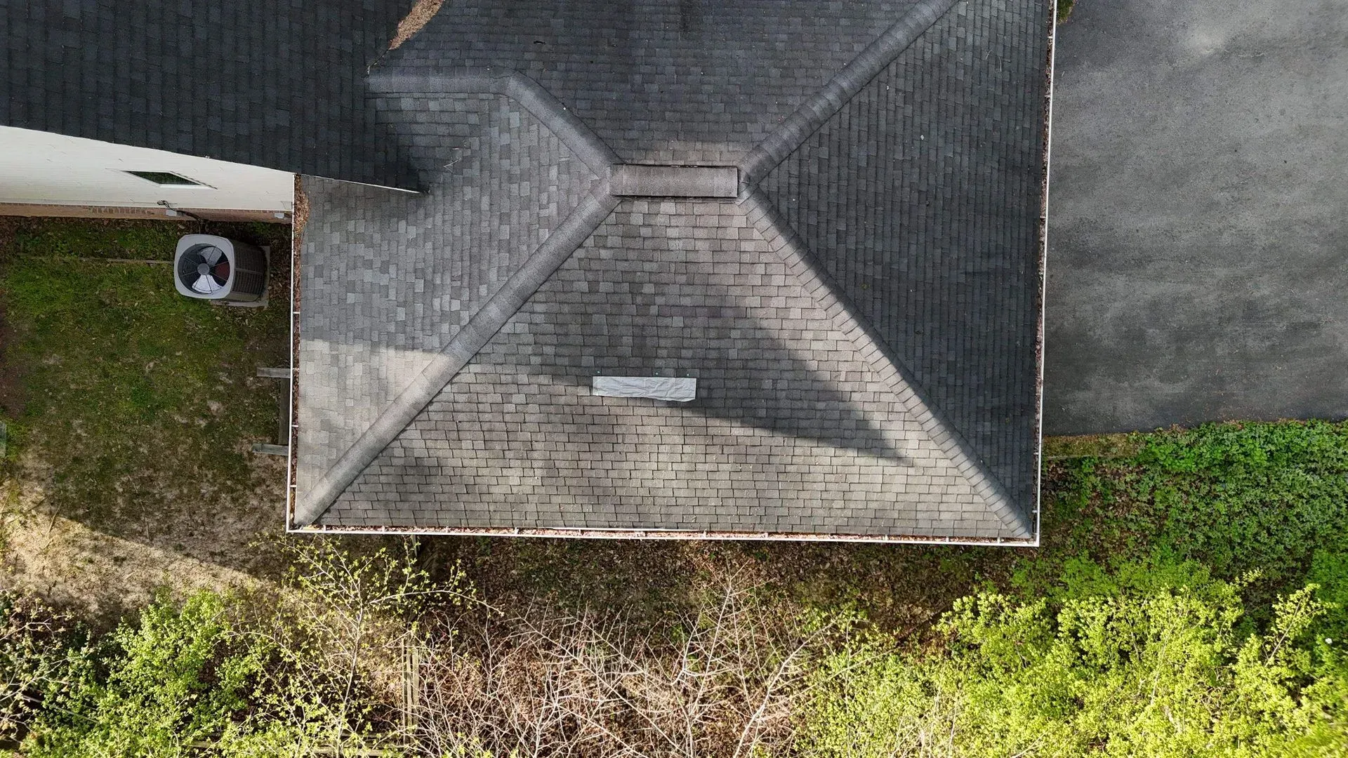 Aerial view of a gray roof with a small vent, bordered by grass and trees.