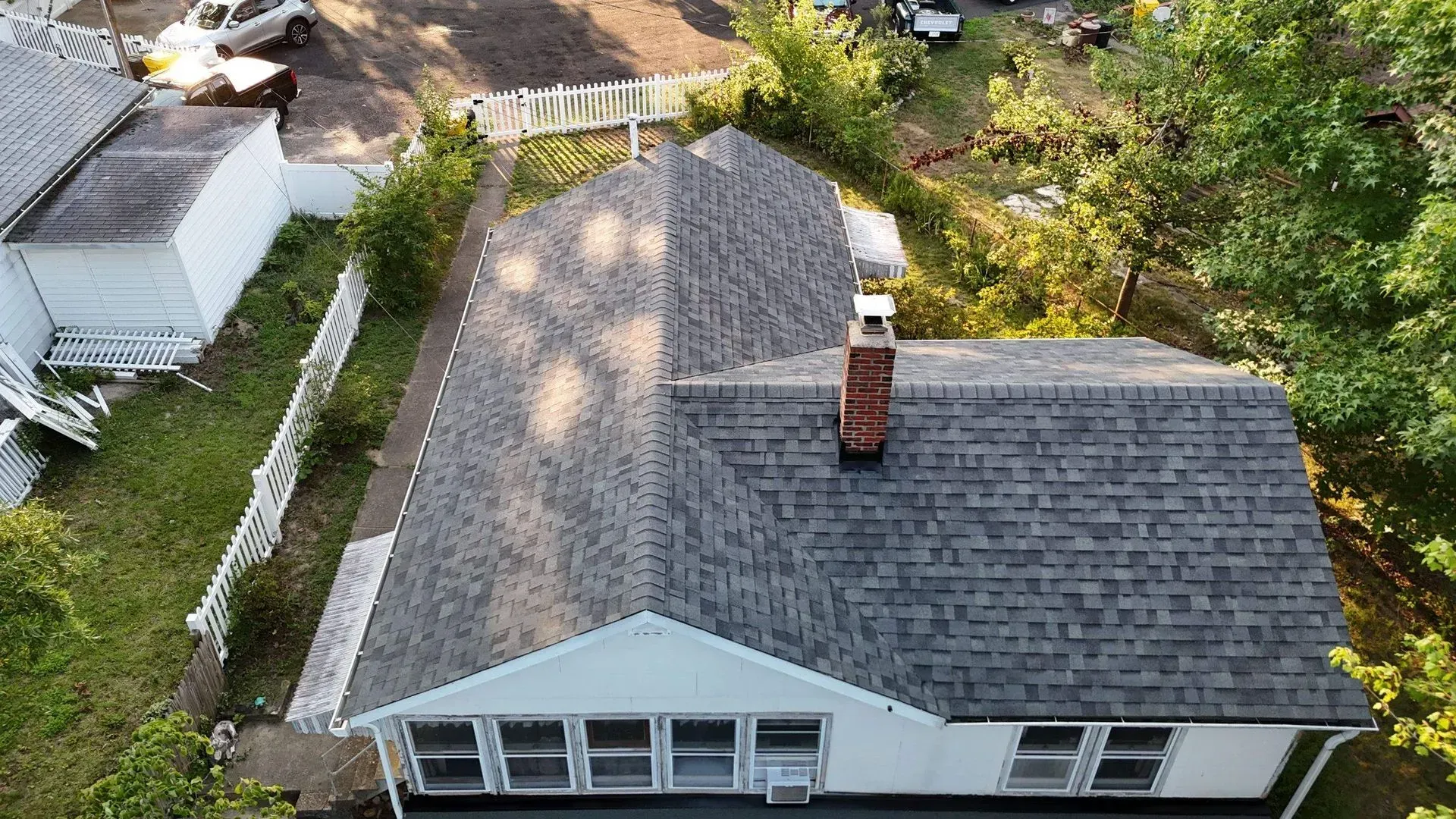 Aerial view of a gray-roofed house with a brick chimney, surrounded by trees and neighboring homes.