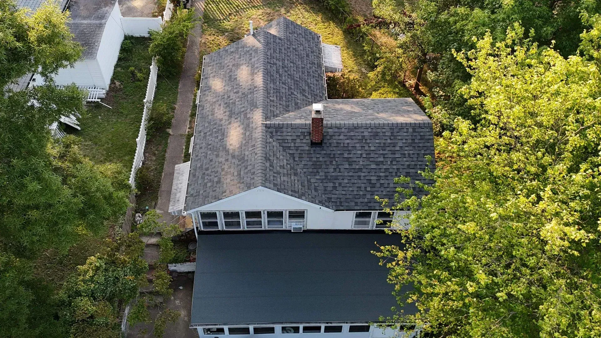 Aerial view of a house with gray shingles, a dark roof, and trees surrounding it