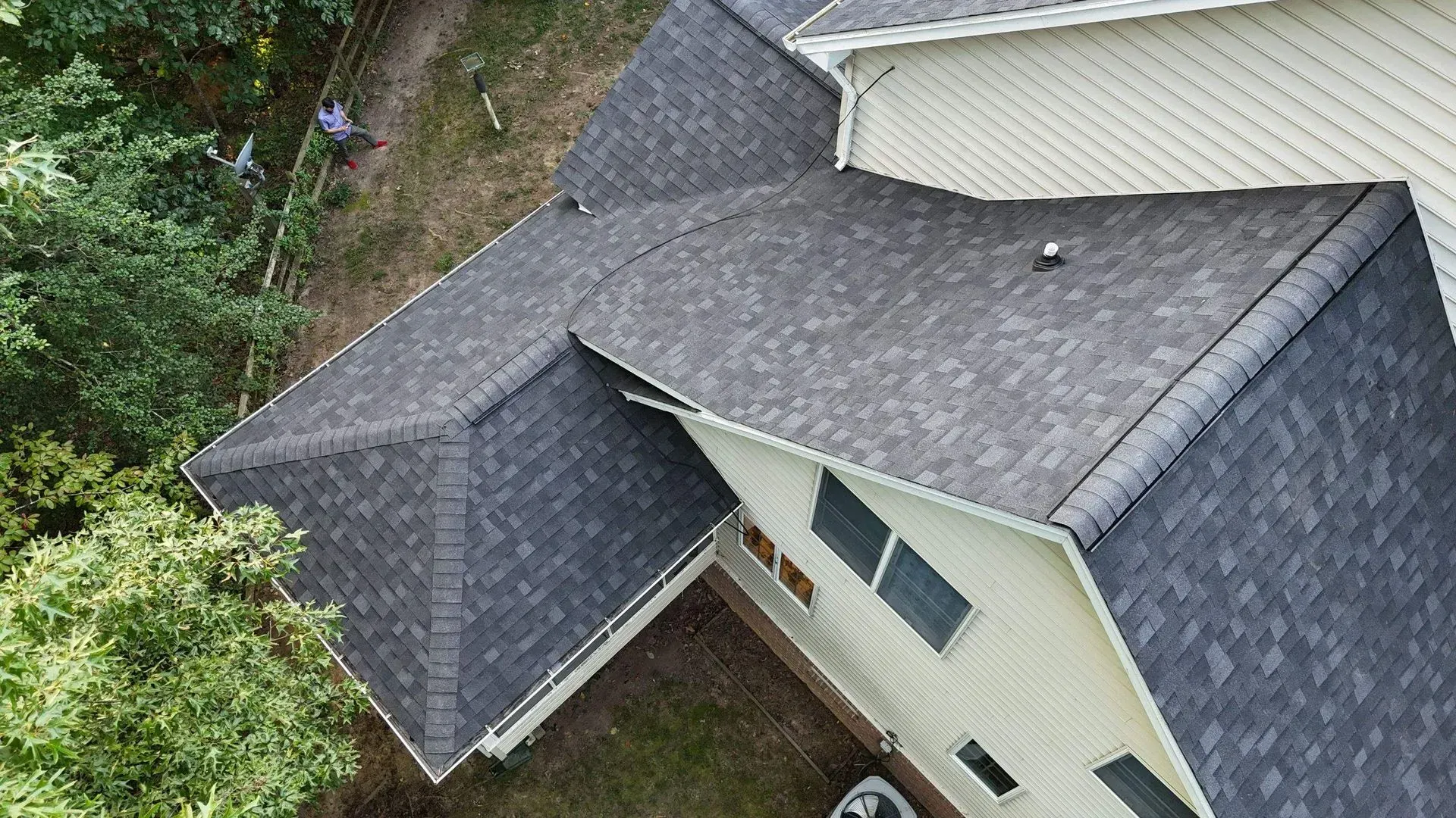 Aerial view of a house roof with dark shingles and a small dormer above trees and siding.