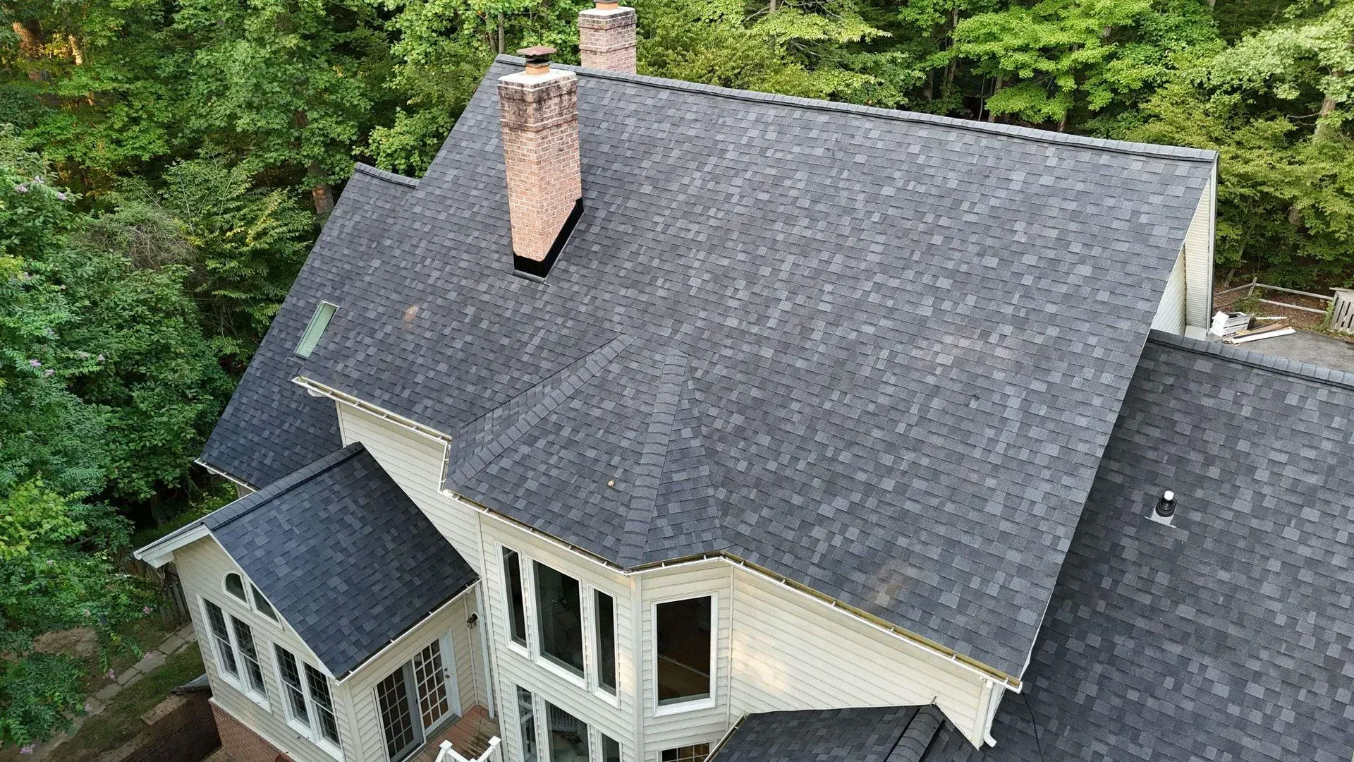 Aerial view of a white house with a dark gray roof and chimney, surrounded by dense green trees.