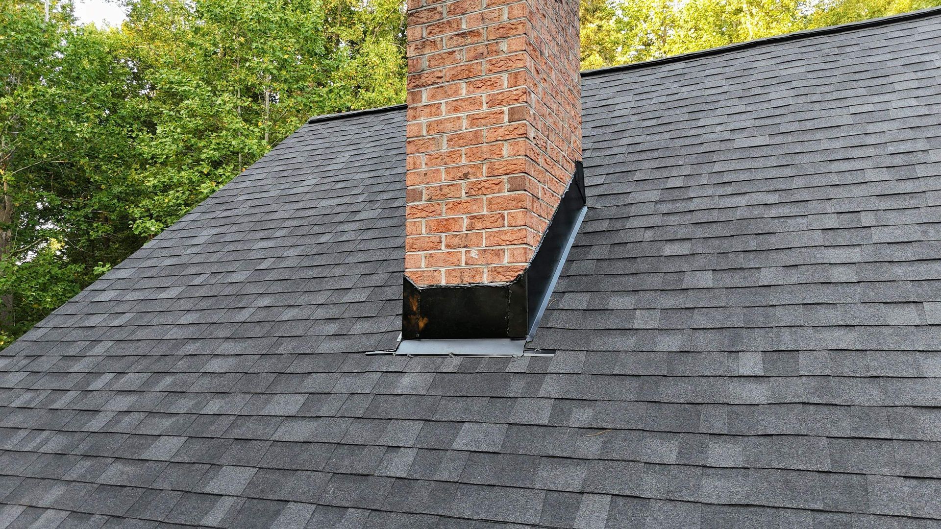 Gray shingled roof with a brick chimney and black flashing, viewed from above with trees in the background