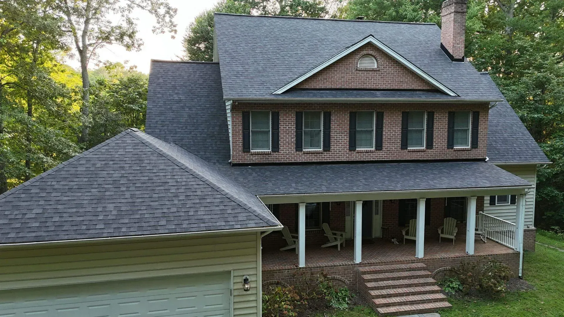 Two-story suburban house with a porch, gray shingled roof, and attached garage amid trees