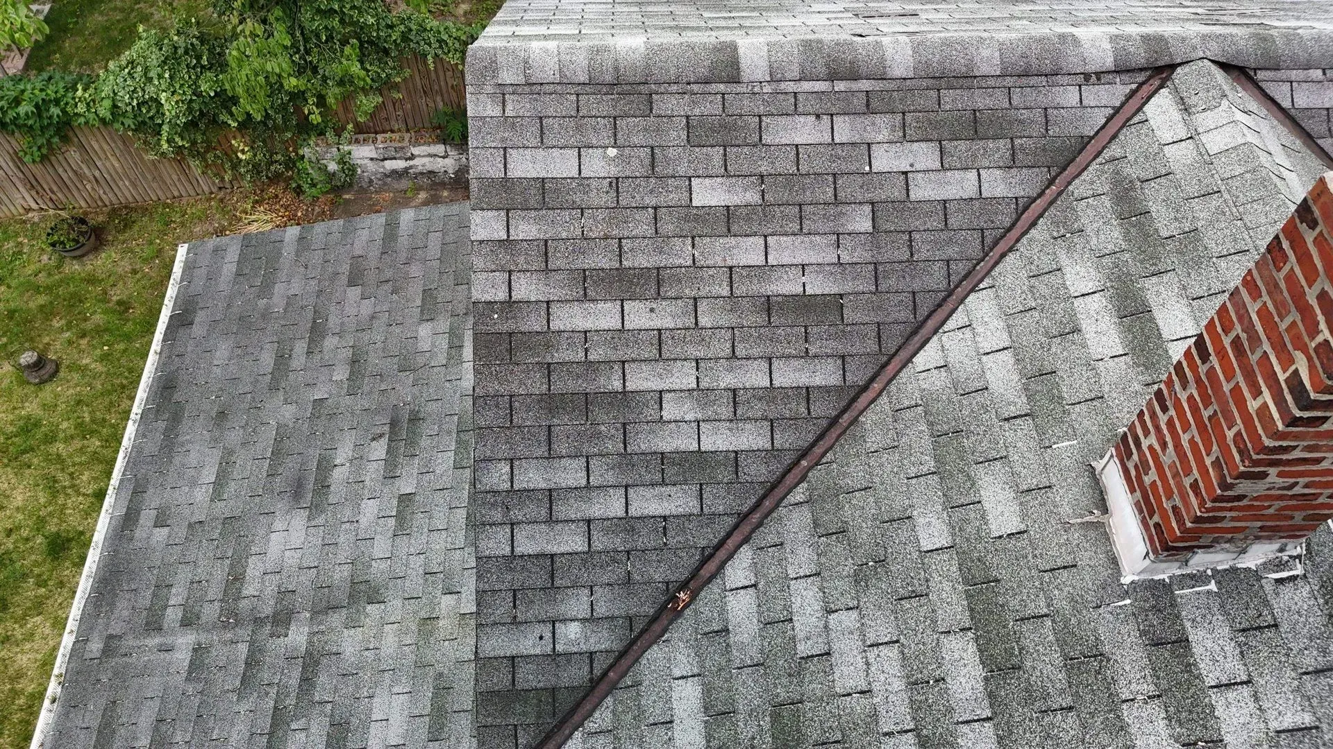 Aerial view of a weathered shingled roof with a brick chimney and adjoining flat roof sections.