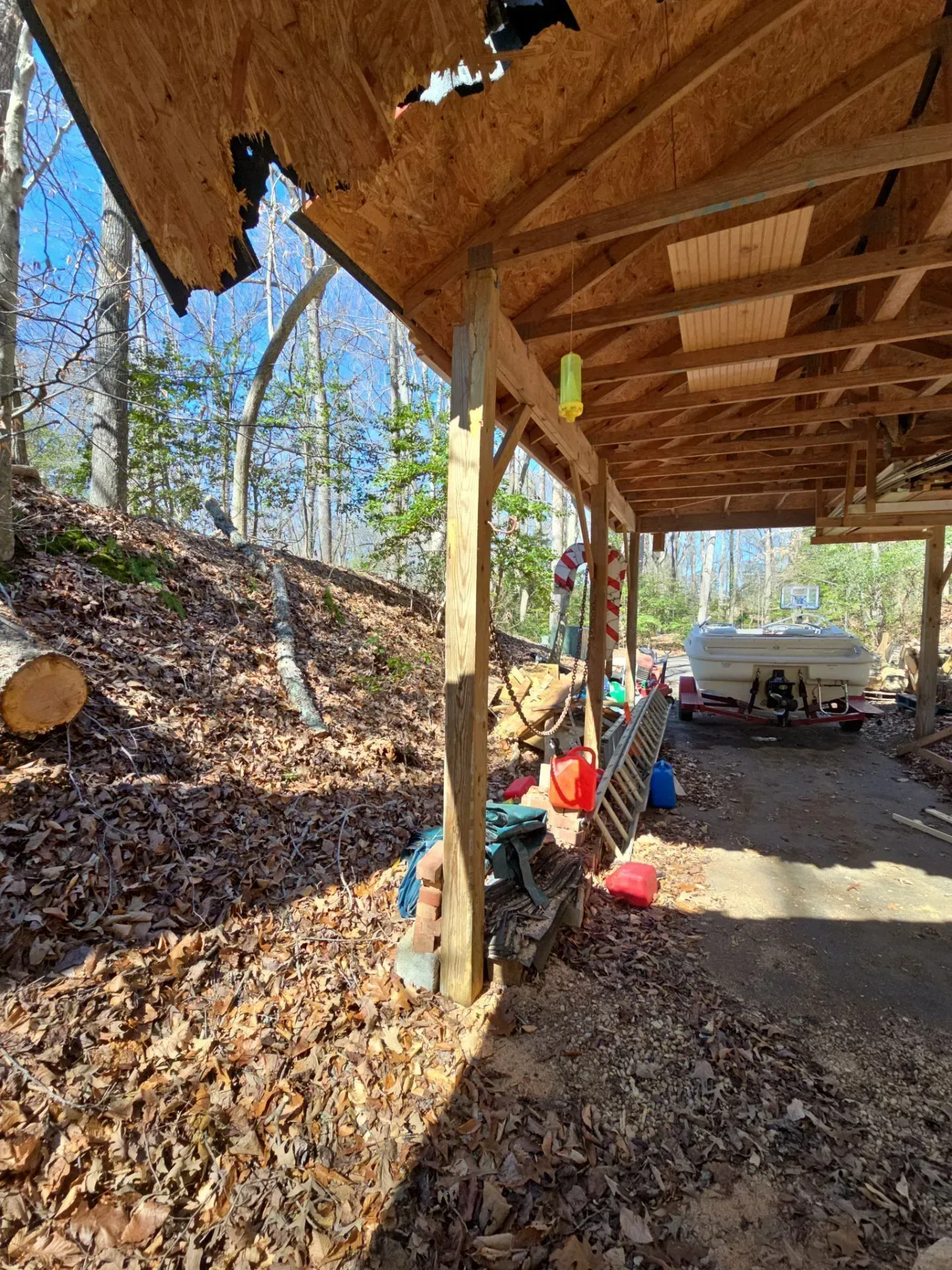 Wooded driveway under a timber carport, with tools, leaves, and a white boat parked at the far end.