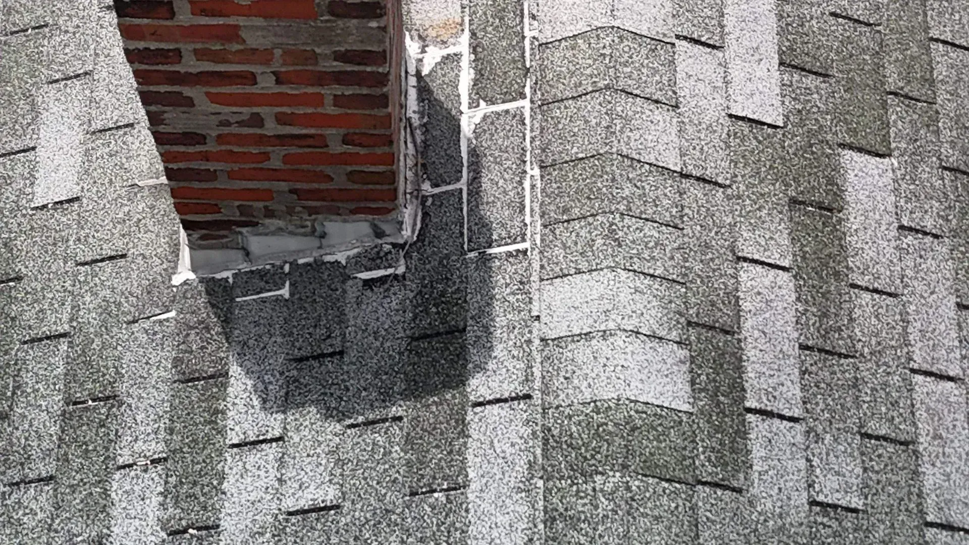 Damaged gray shingle roof with a broken opening exposing red brick and metal flashing
