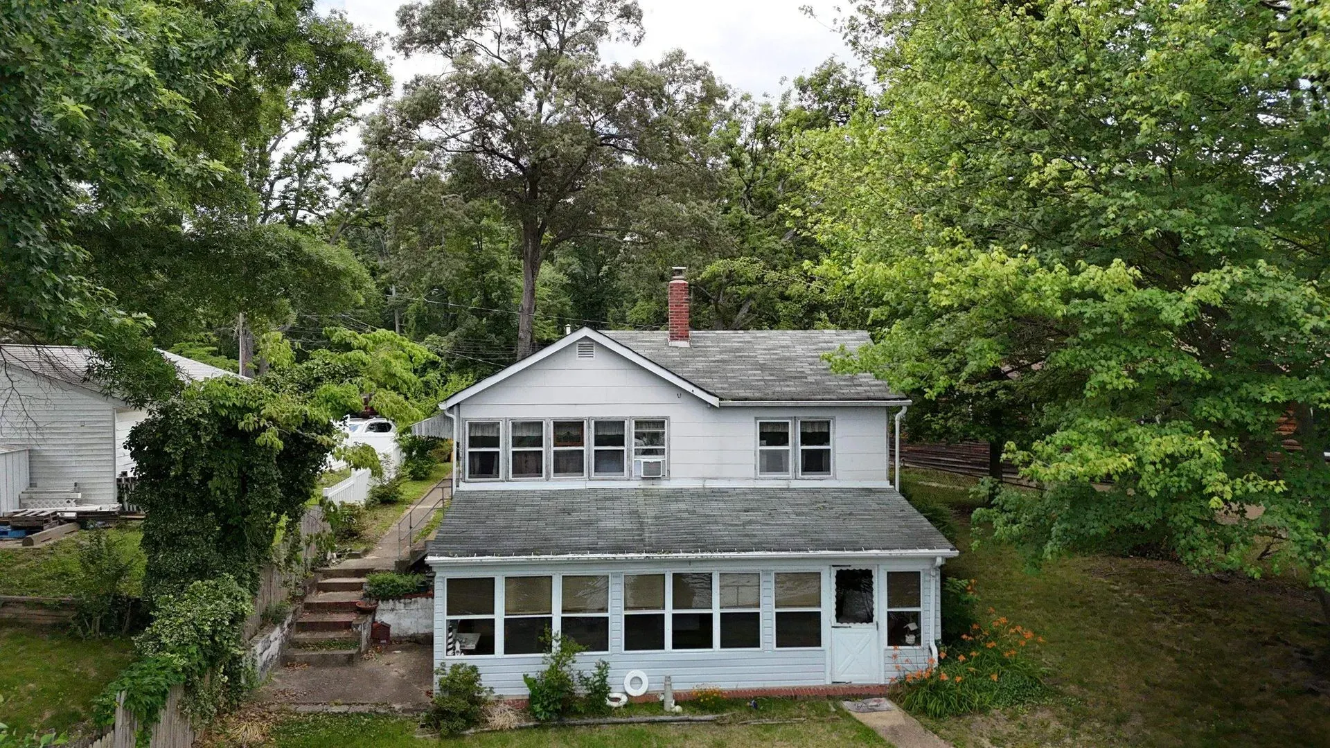 White two-story house with a screened porch, surrounded by trees and greenery on a hillside.