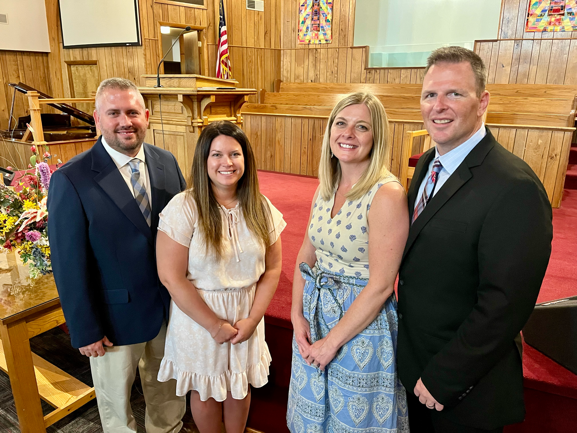 Four formally dressed people pose in a church. Two men wear suits. Two women wear dresses.