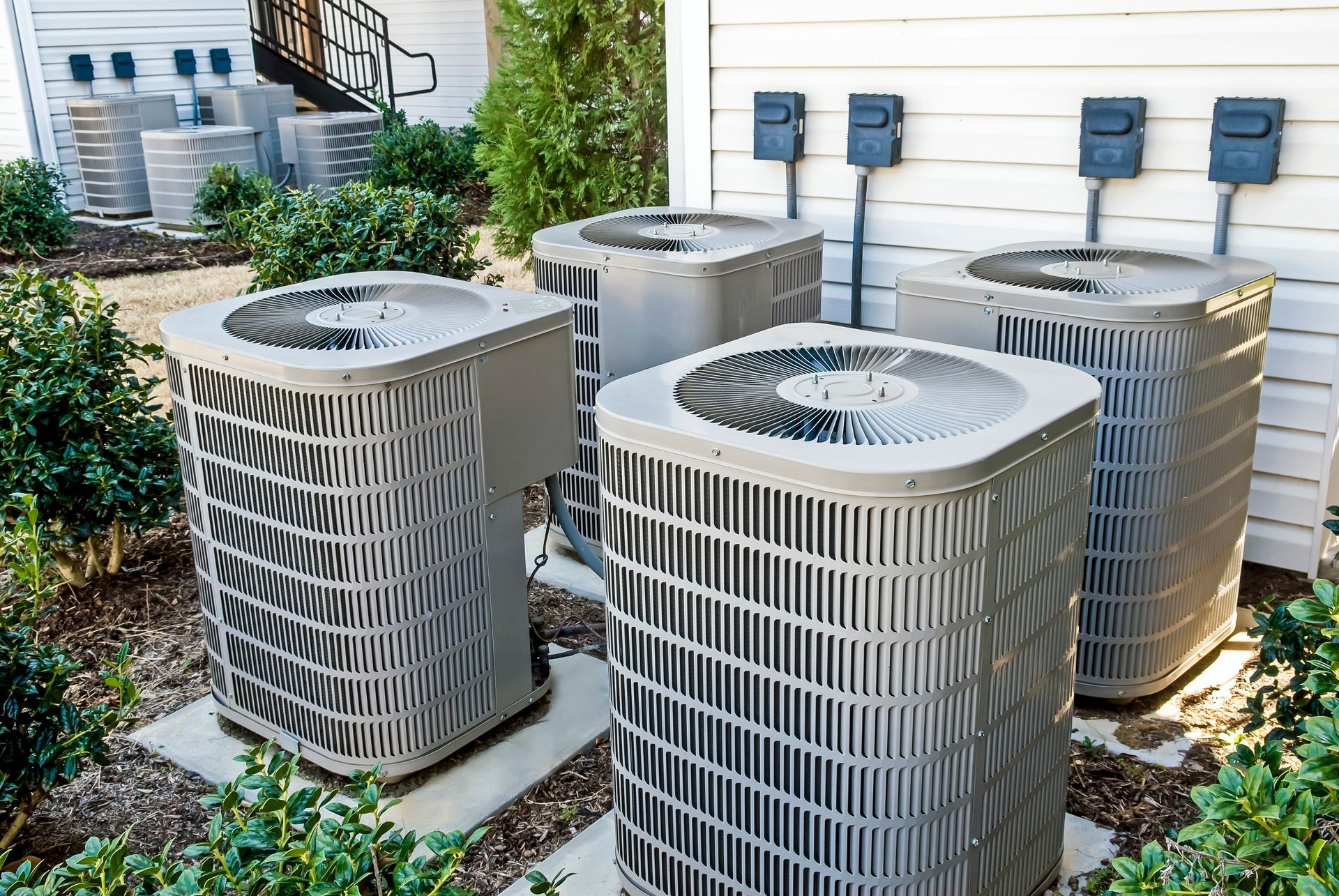 Air conditioning units lined up outdoors against a white wall with electrical connections.