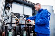 A technician in blue overalls is repairing machinery, using a red device, outdoors.