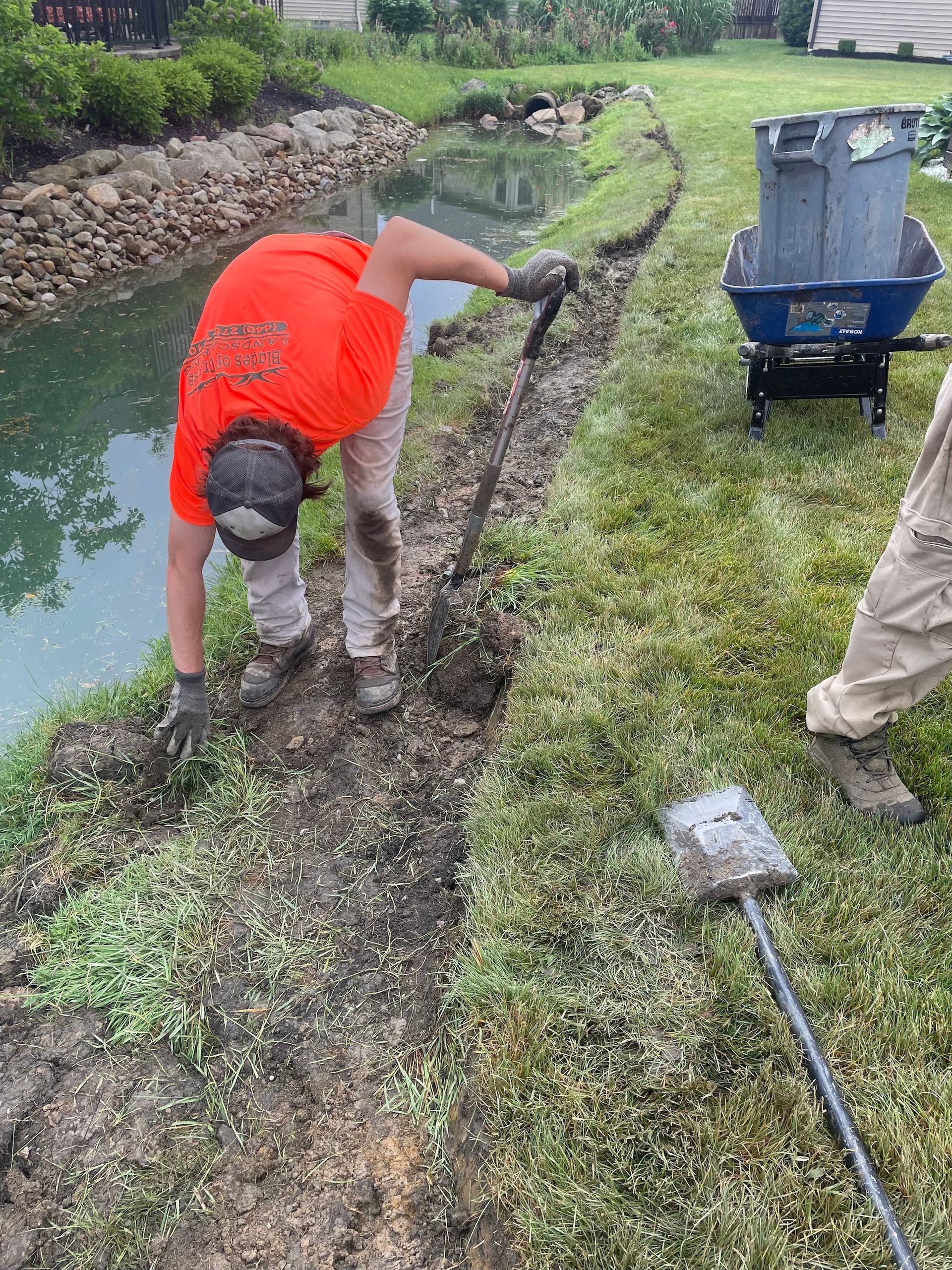 A man in an orange shirt is digging in the dirt next to a wheelbarrow.
