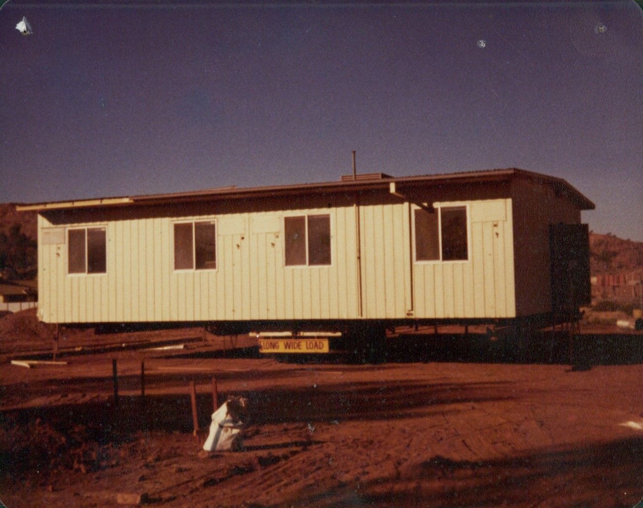 A rectangular, off-white mobile building sits on a trailer in a dusty, sunlit outdoor lot.