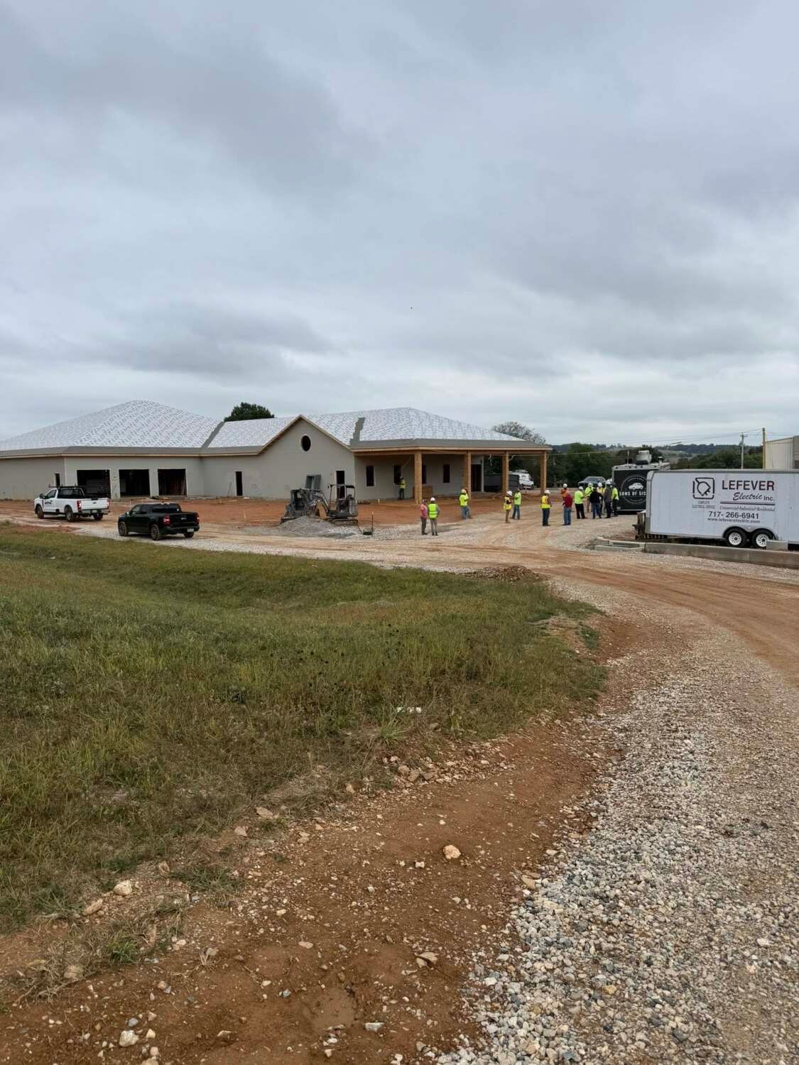 Building construction site; workers near a partially built building with a gravel road. Overcast sky.