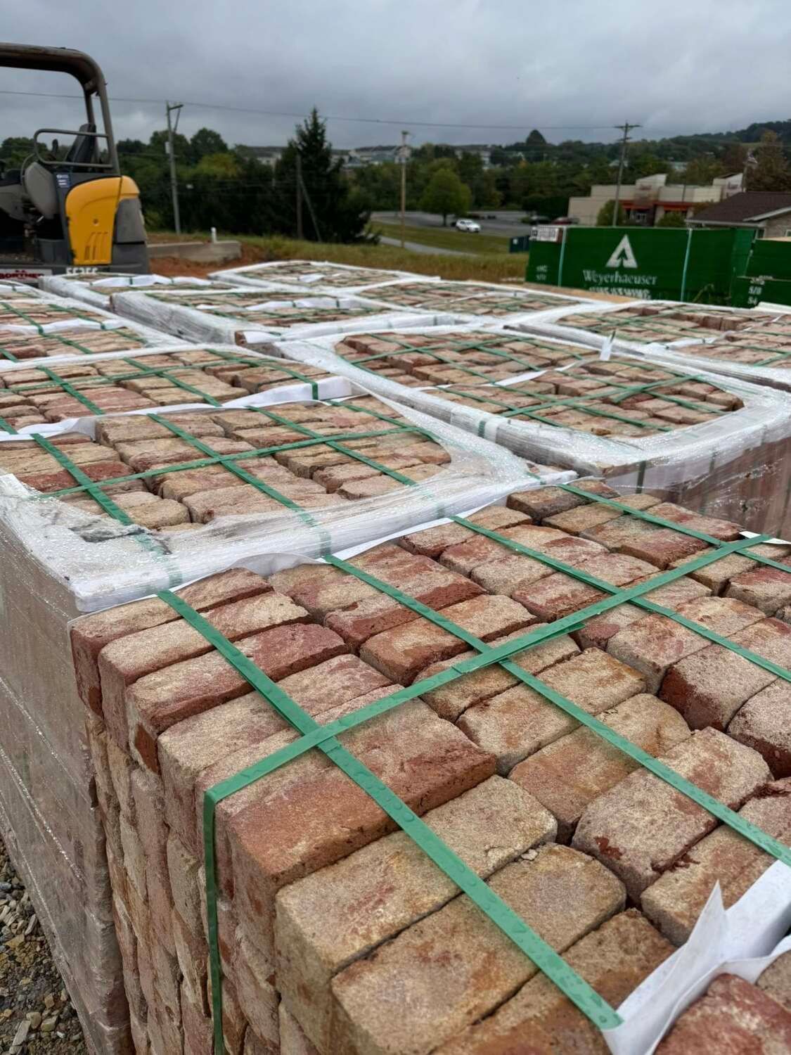 Pallets of red-brown bricks, secured with green straps, outdoors under a cloudy sky.