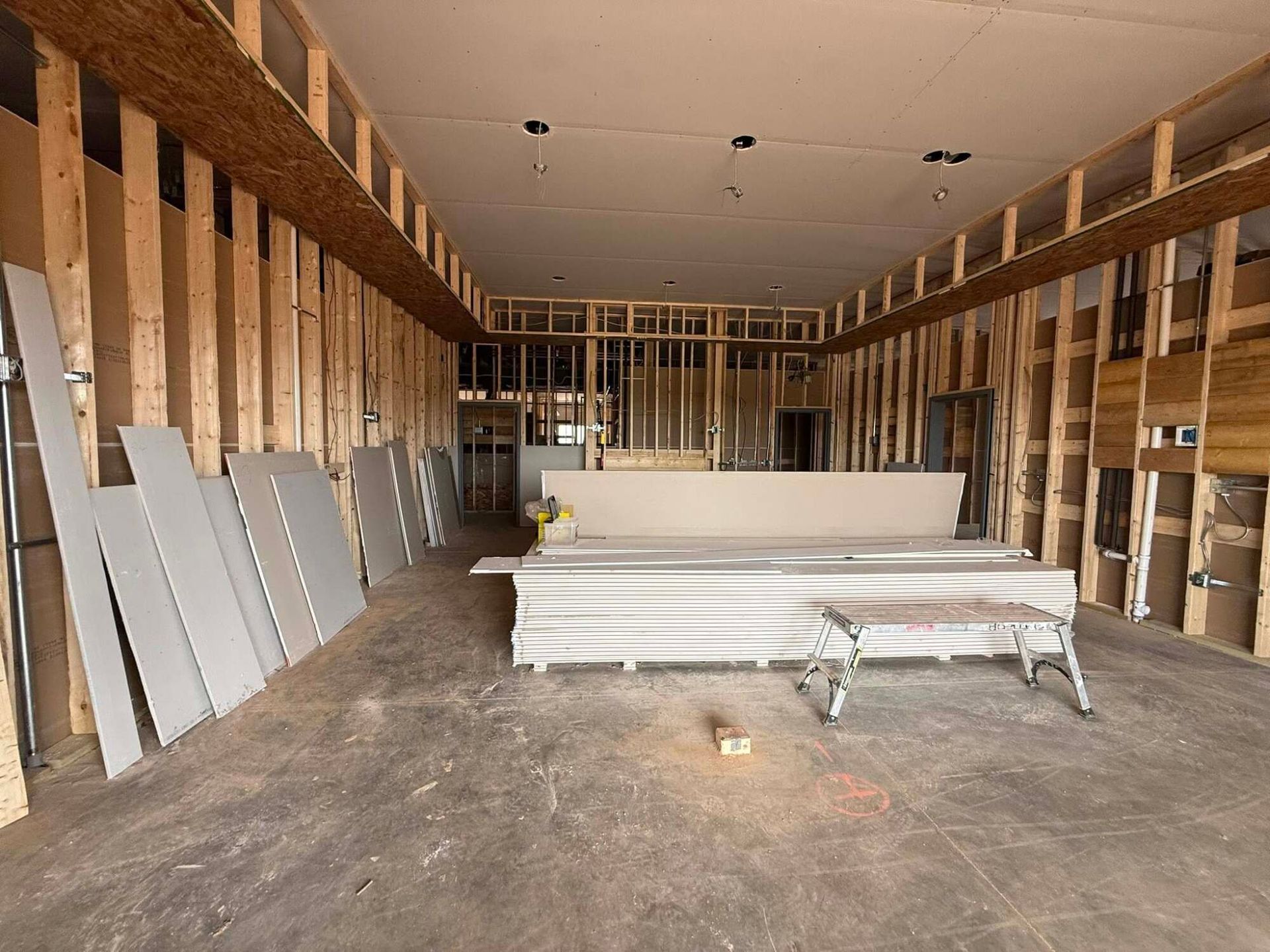 Interior of a building under construction, showing exposed wooden framing, drywall, and unfinished concrete floor.