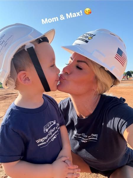 Mother and child wearing hard hats share a kiss at an outdoor construction site together joyfully