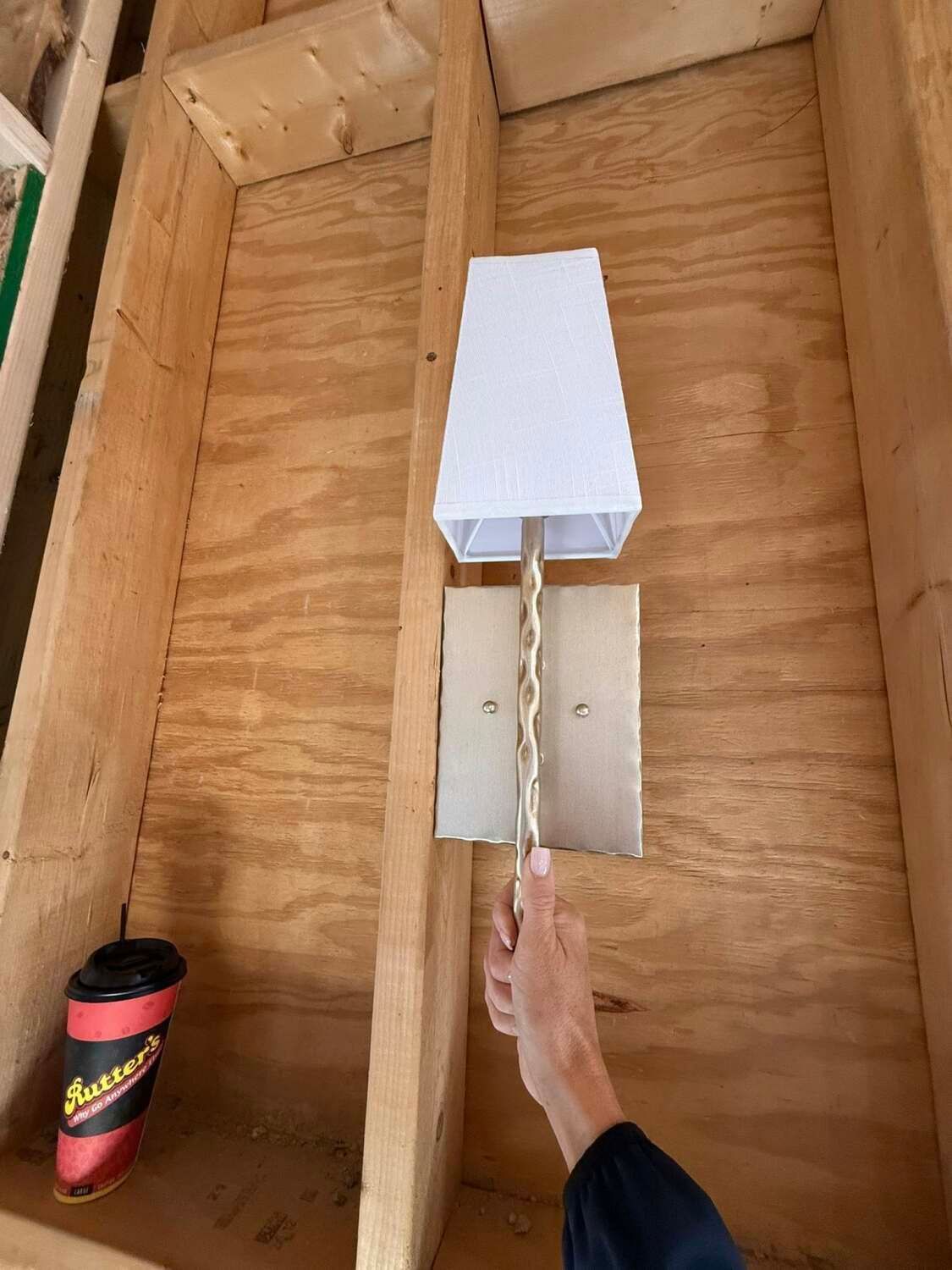 A hand holding a wall lamp with a white lampshade and gold chain, mounted on a wood wall.