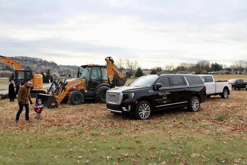 Black SUV and construction equipment parked near a grassy field; two people walk in the foreground.
