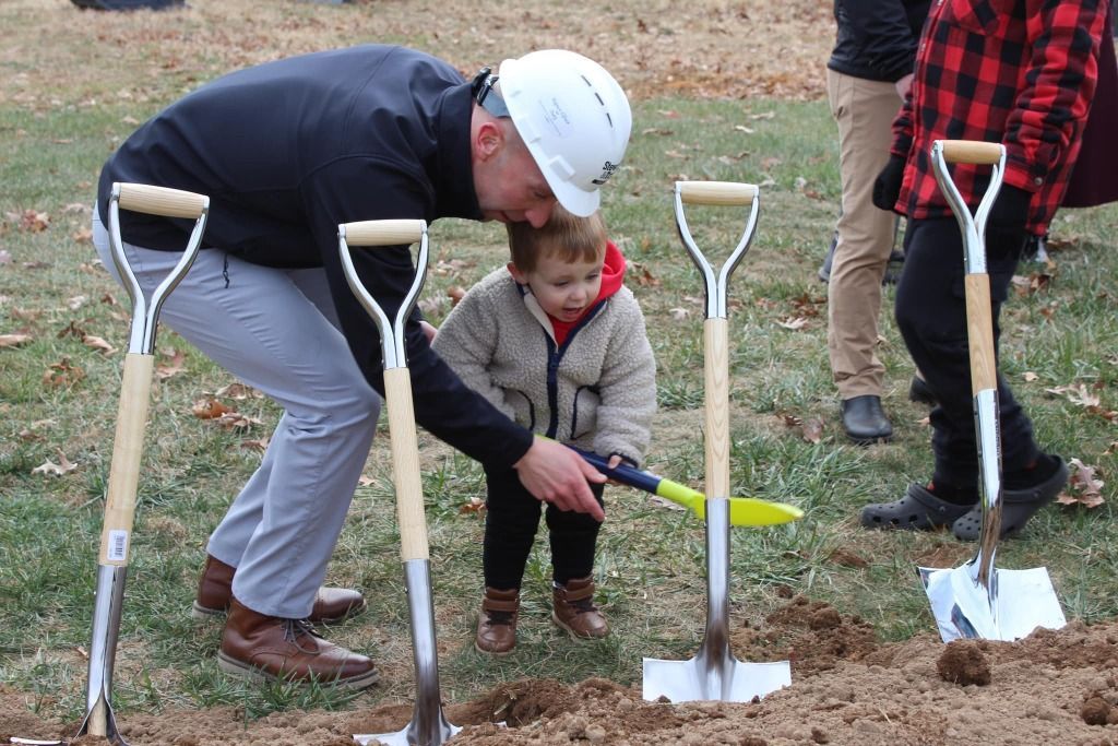 Man in hard hat helps child dig with shovel at a groundbreaking ceremony, several shovels in the ground.
