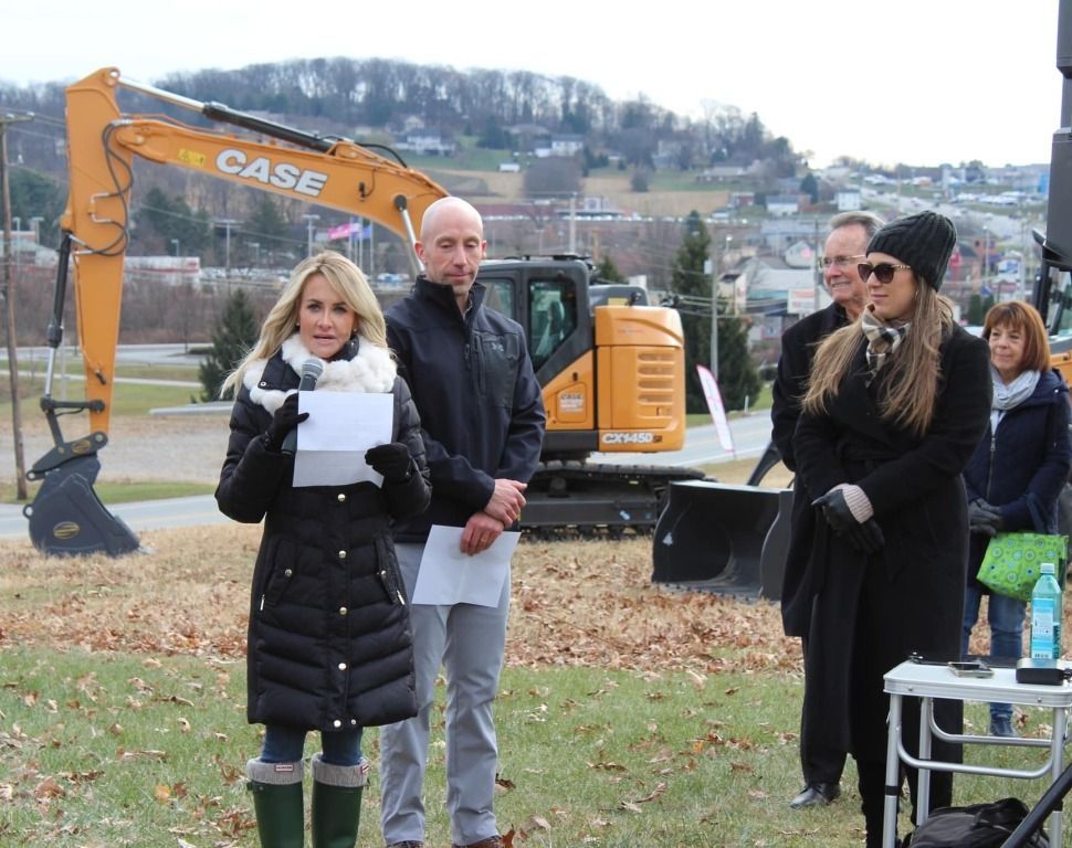People at an outdoor event with an excavator. One woman speaks, holding papers. Other people stand nearby.