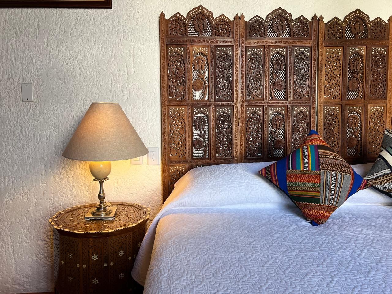 Dormitorio con cabecera de madera ornamentada, mesa auxiliar, lámpara, obras de arte y ropa de cama blanca.