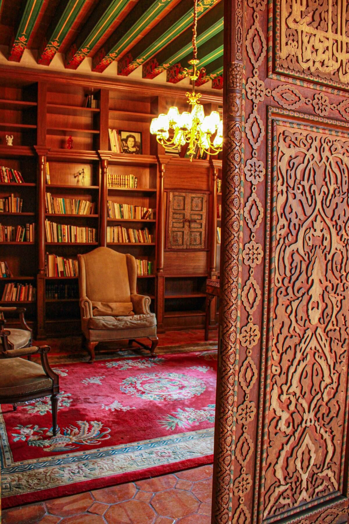 Una biblioteca con estanterías de madera, alfombra roja, sillón, lámpara de araña y puerta de madera tallada.