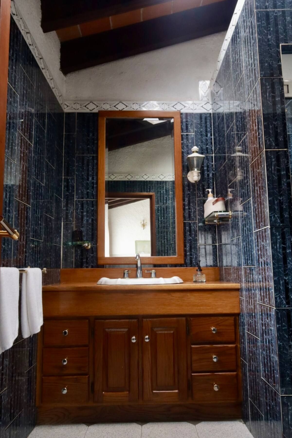 Tocador de madera en un baño con paredes de azulejos azul oscuro, espejo y luz natural.