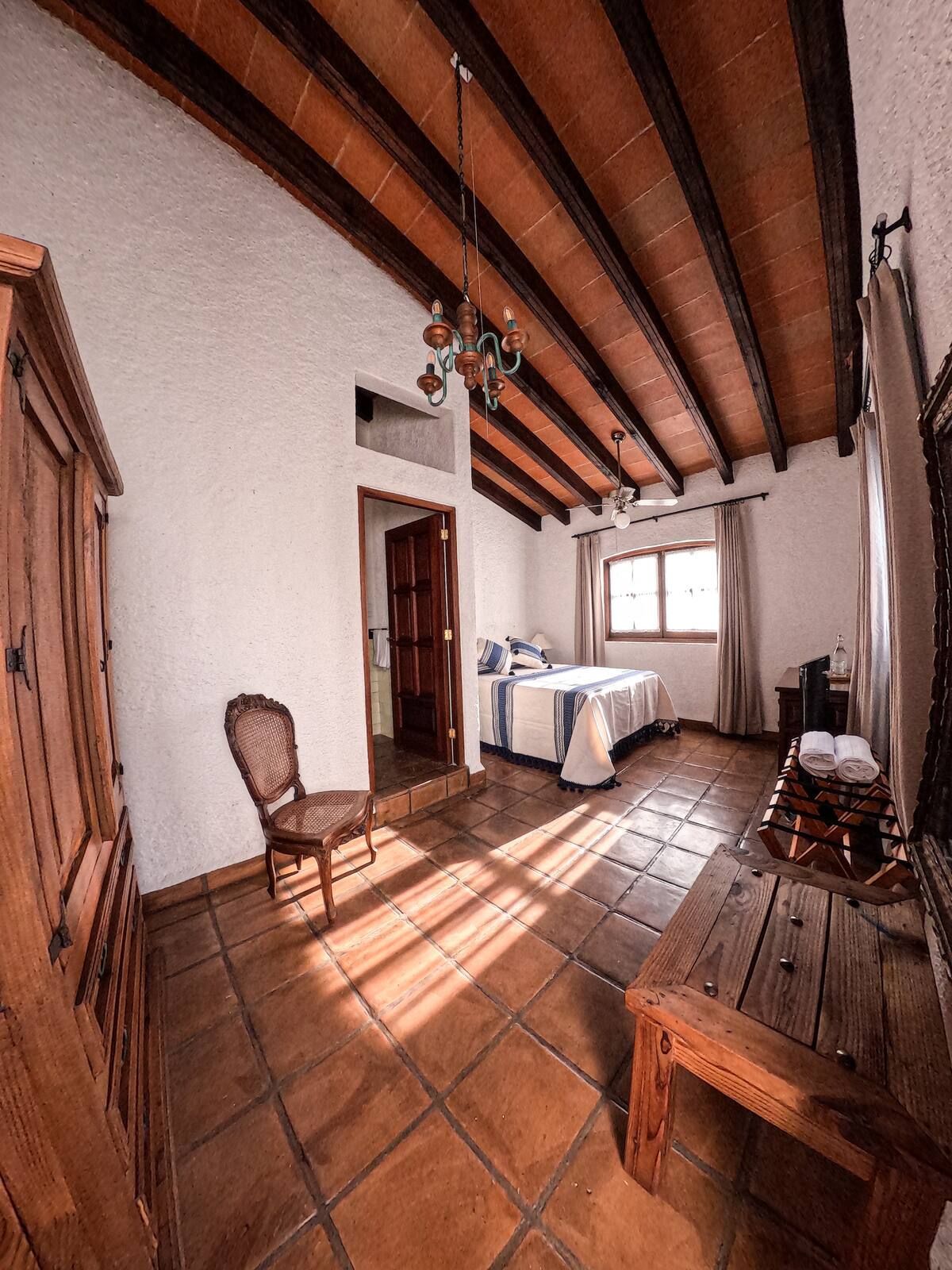 Dormitorio con suelo de terracota, vigas de madera y muebles antiguos. La luz del sol entra a raudales.