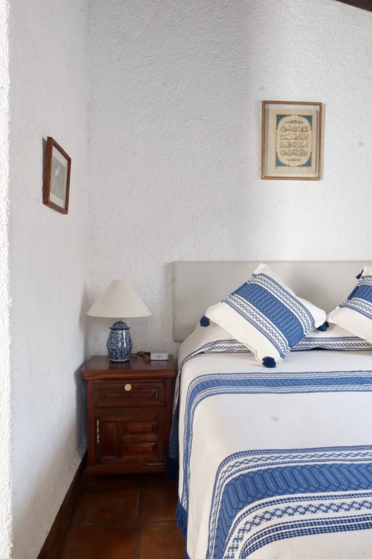 Dormitorio con paredes texturizadas blancas, cama con detalles azules, mesita de noche con lámpara y obras de arte.