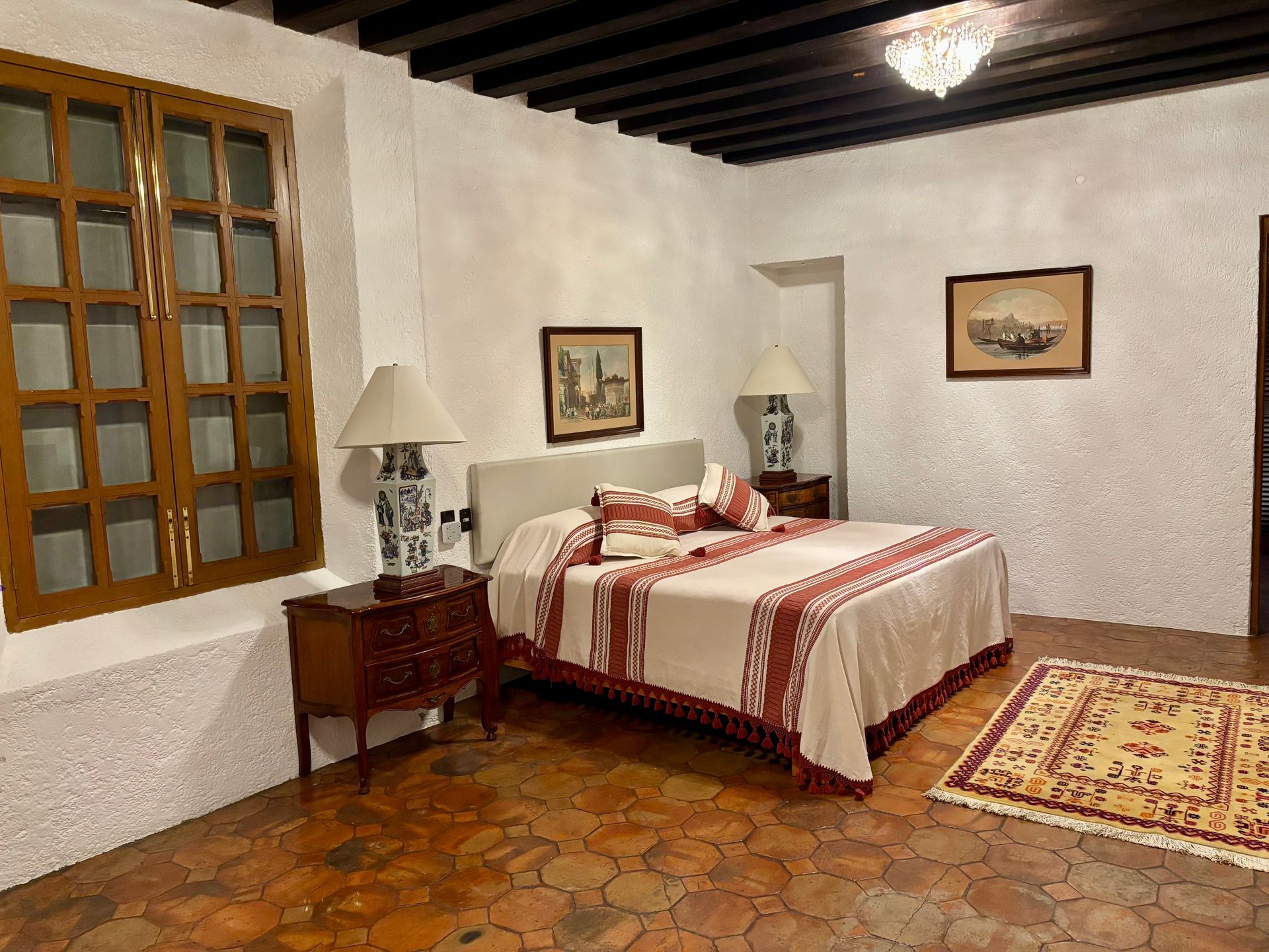 Dormitorio con cama, mesitas de noche, alfombra y cuadros, en una habitación con ventana y techo de madera oscura.