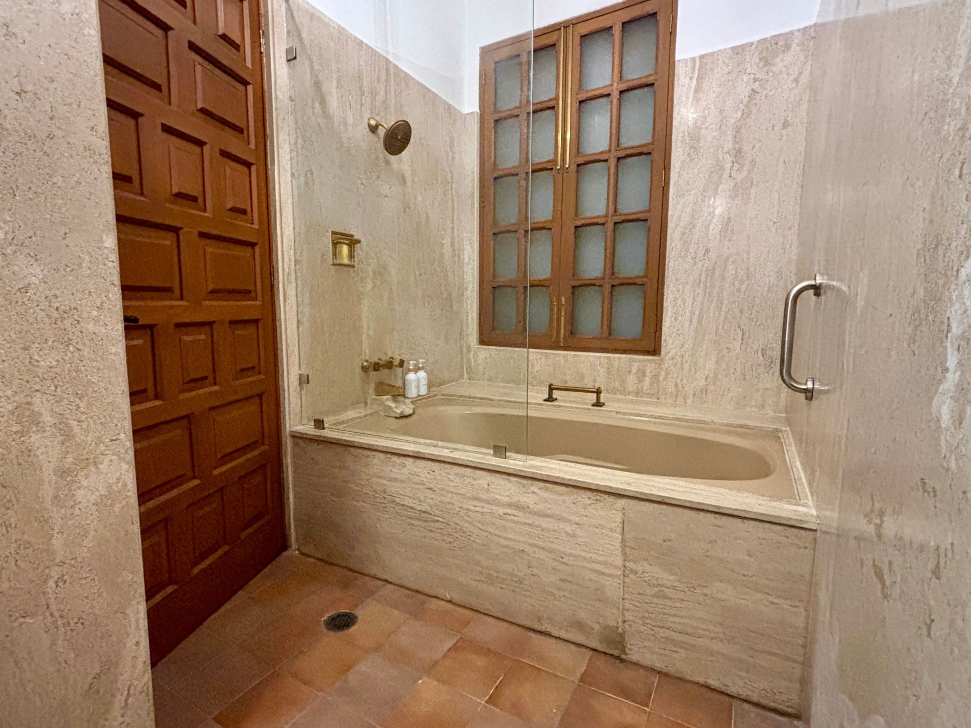Baño con bañera, puerta de madera y ventana. Paredes y suelo de azulejos beige.