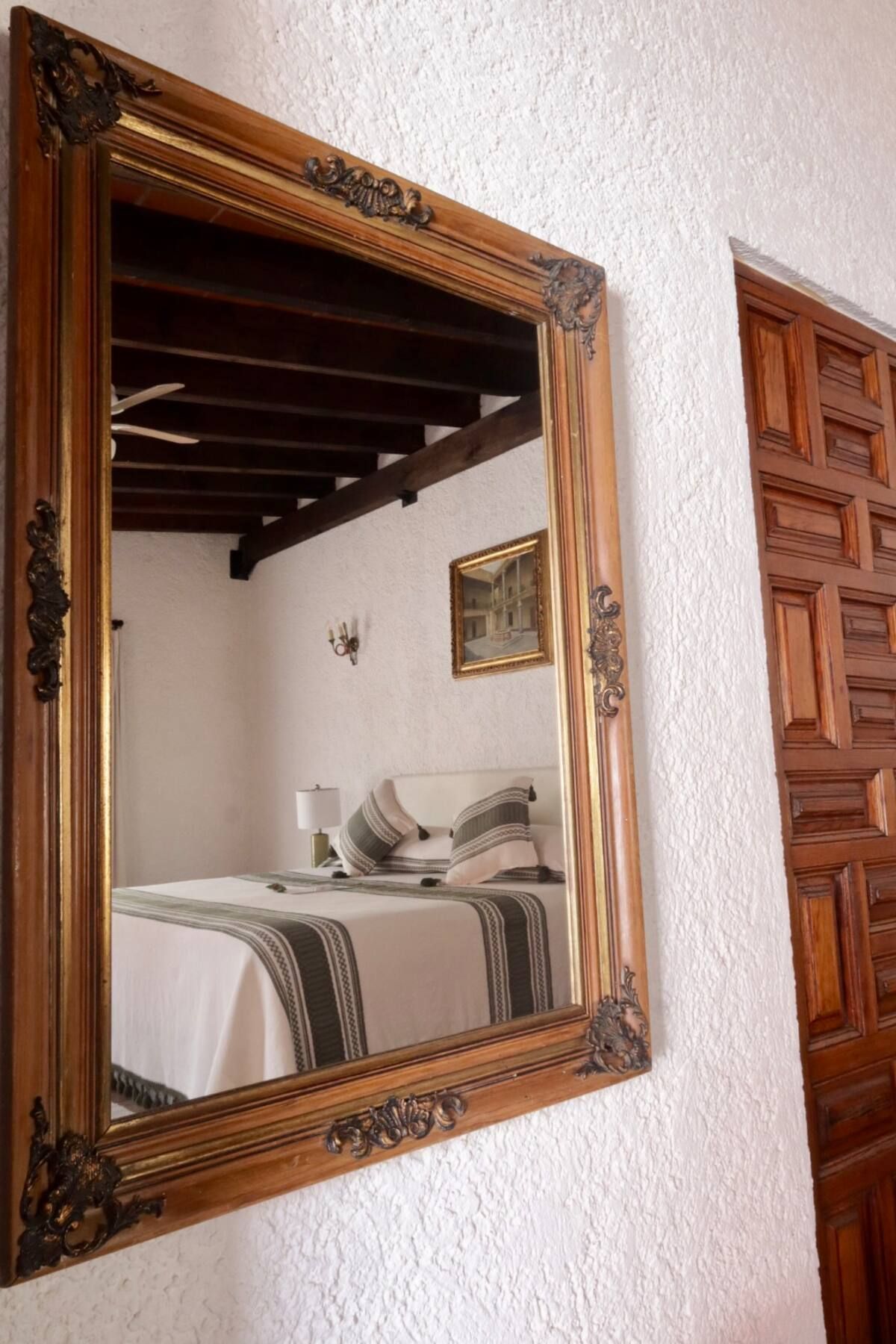 Espejo sobre pared blanca que refleja un dormitorio con cama y vigas de madera.