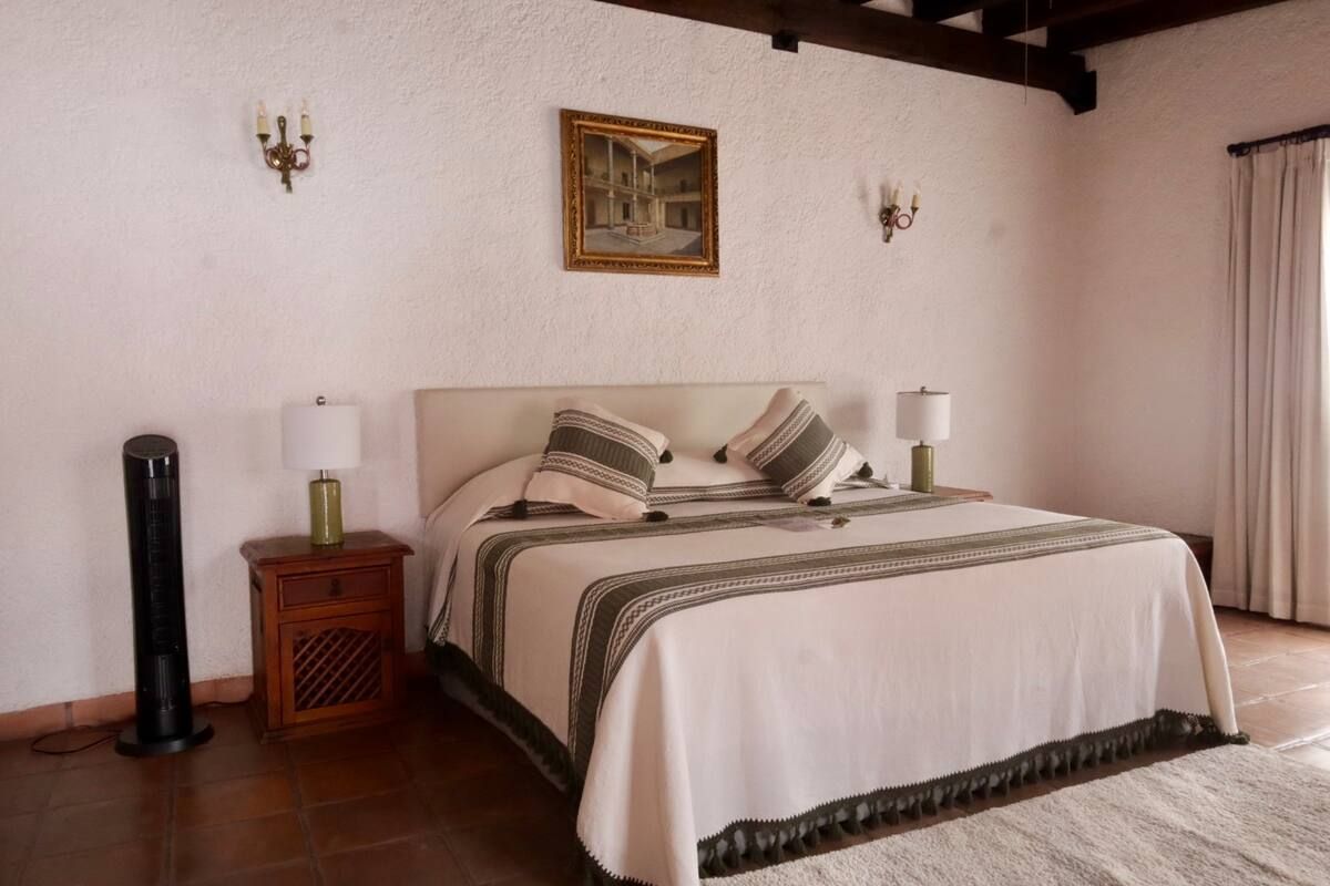Dormitorio con cama, mesitas de noche, lámparas, obras de arte, ventilador y alfombra. Paredes blancas y suelo de madera.