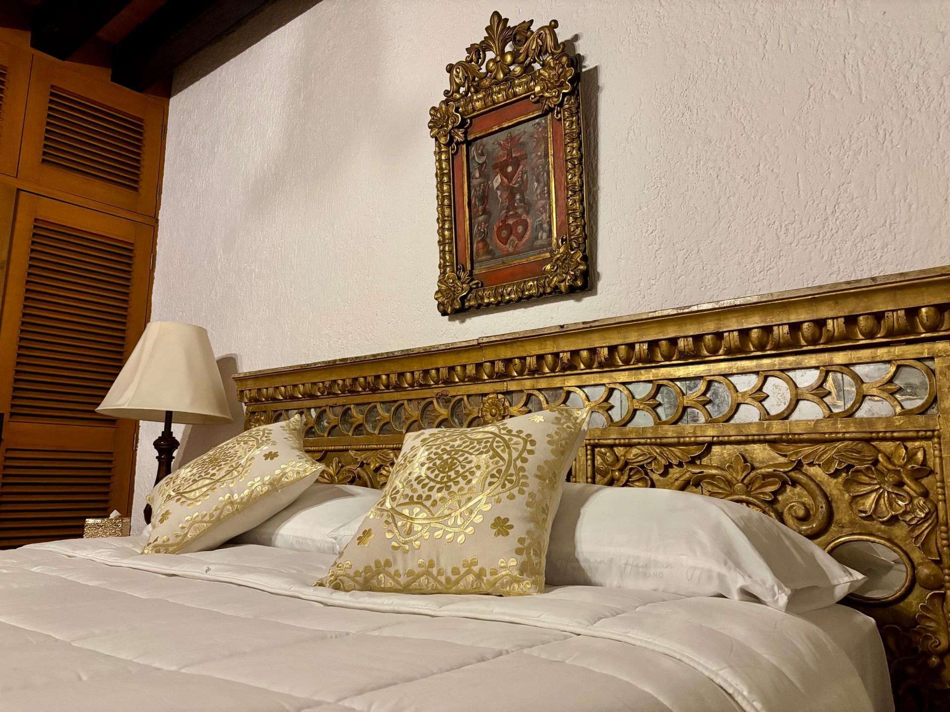 Cabecero dorado ornamentado con ropa de cama blanca y almohadas decorativas, debajo de una pintura religiosa.