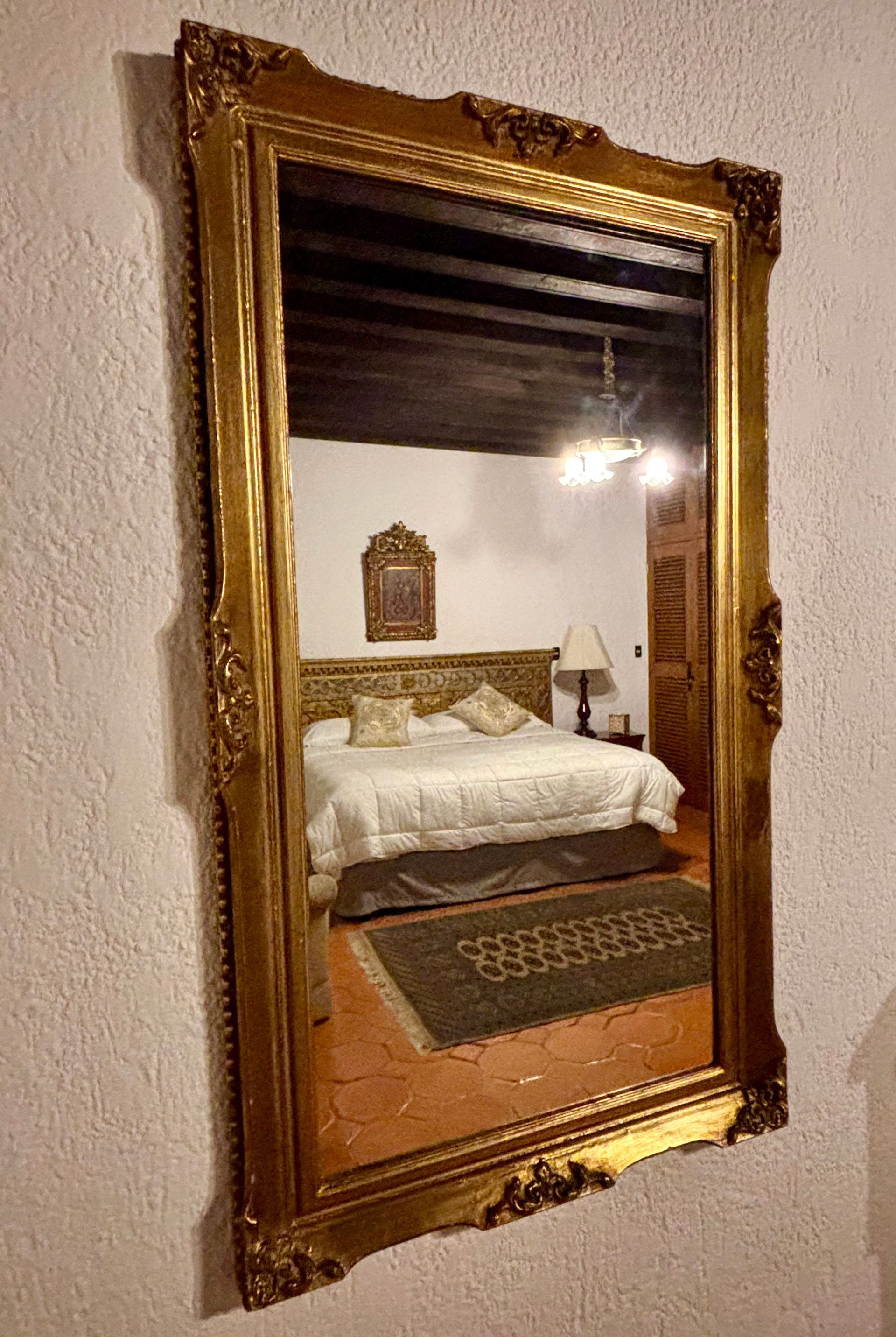 Espejo adornado con marco dorado que refleja un dormitorio con una cama, una alfombra y una lámpara de araña.