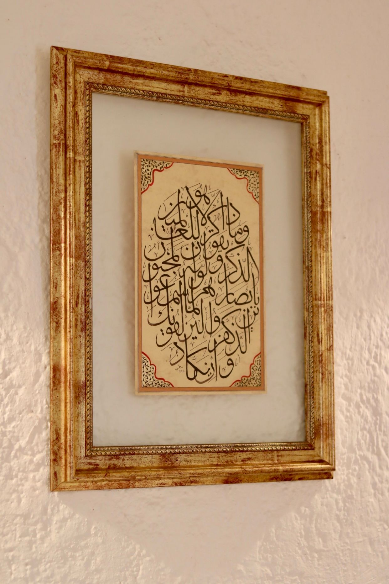 Obra de arte de caligrafía árabe enmarcada en un marco dorado adornado sobre una pared blanca texturizada.