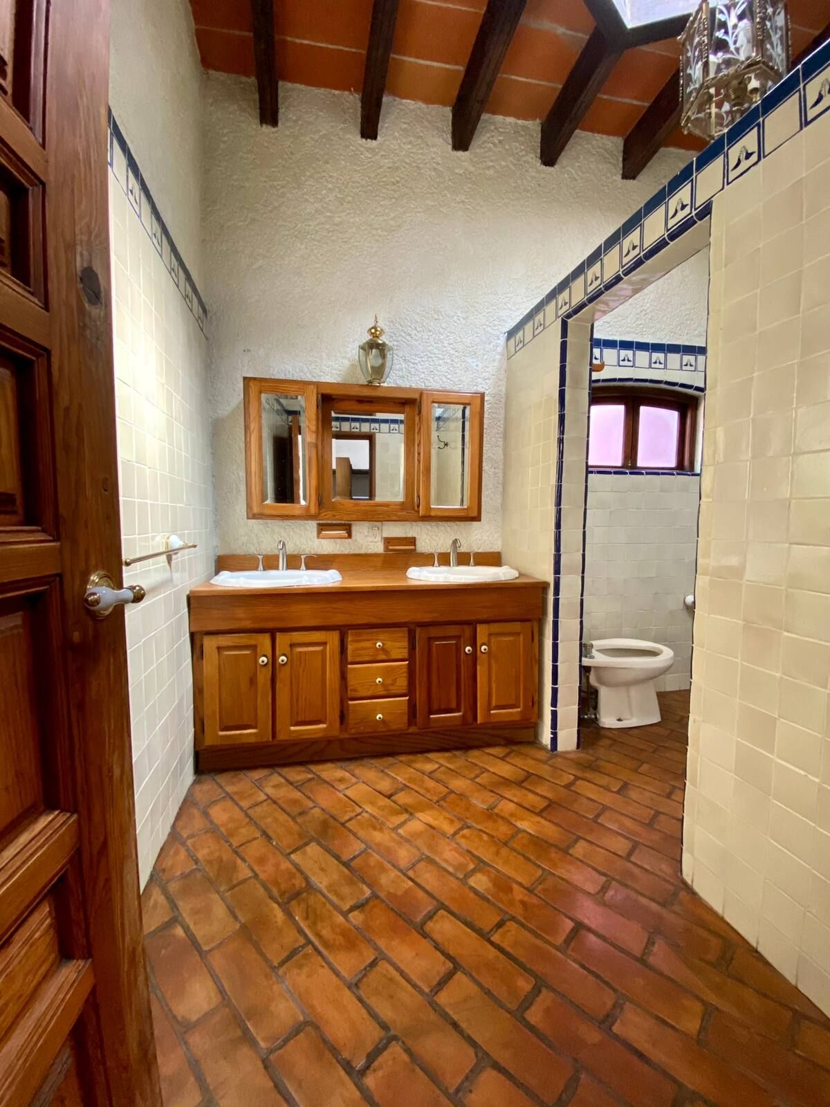 Baño con tocador de madera, piso de ladrillo, azulejos blancos y un inodoro.