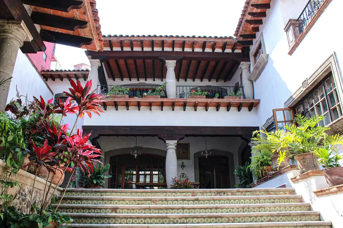 Patio con escalera que conduce a un edificio blanco con arcos y balcones, rodeado de plantas.