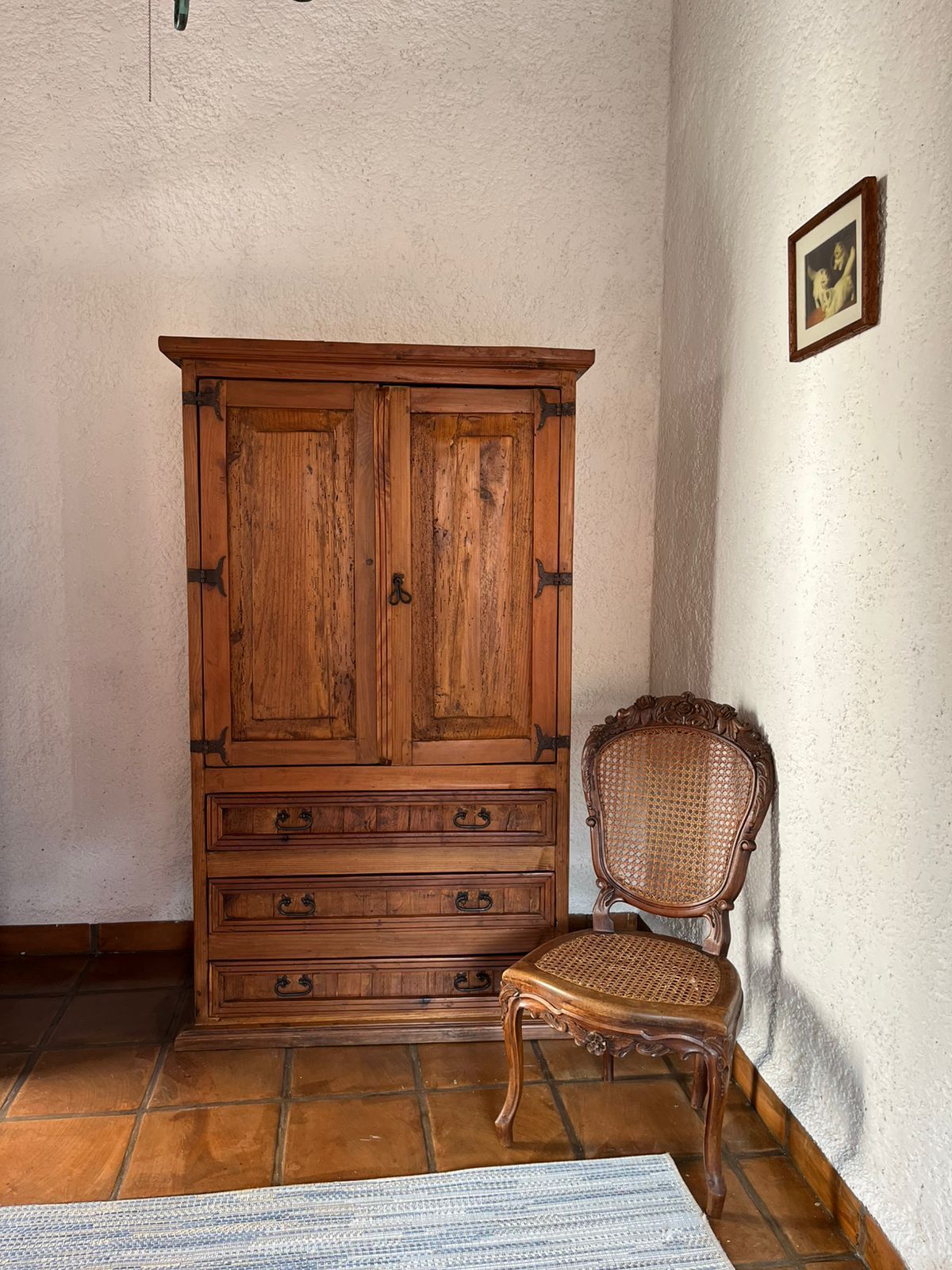 Gabinete de madera, silla tejida, arte enmarcado y piso de baldosas en una habitación con paredes blancas texturizadas.