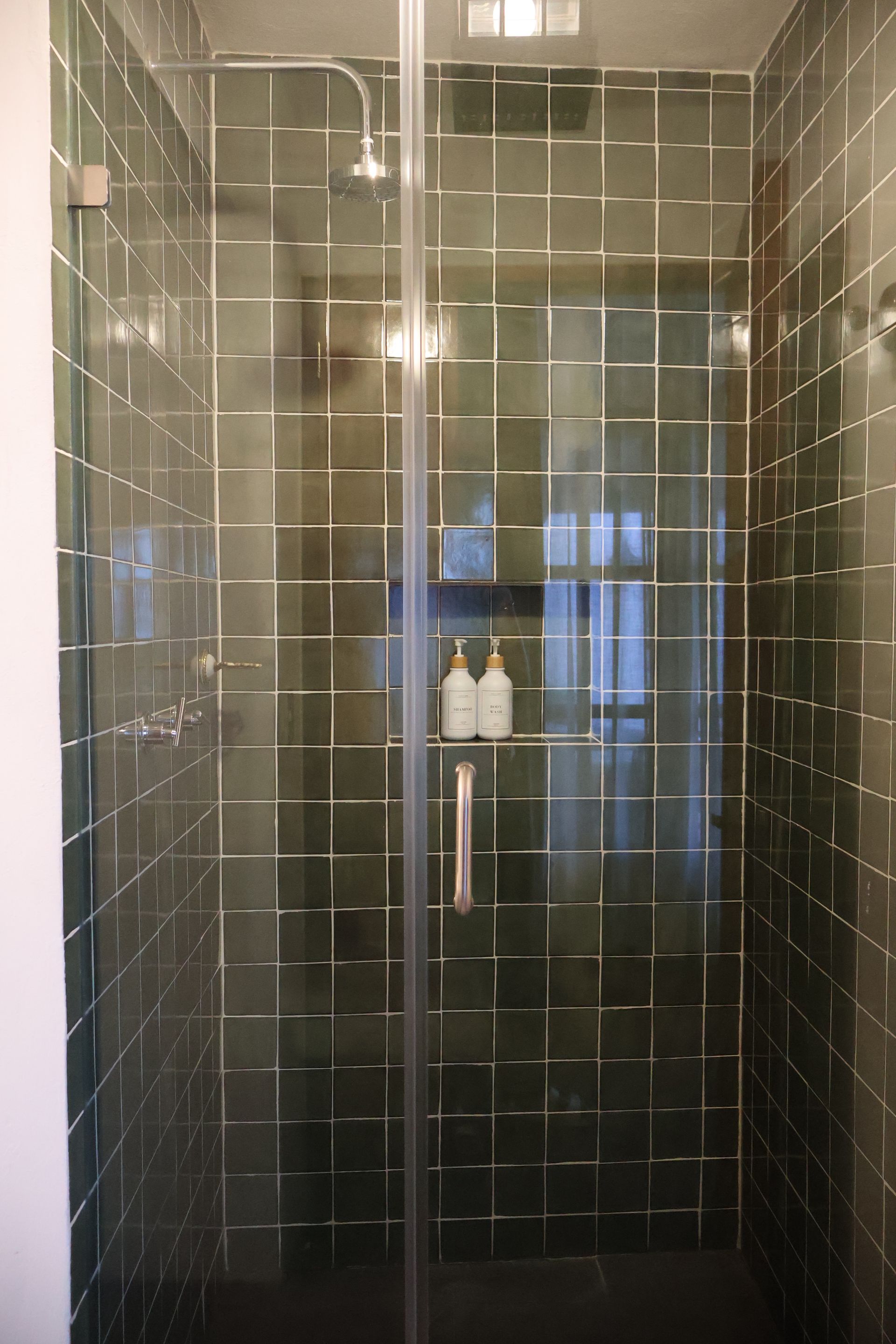 Ducha con azulejos cuadrados verdes y puerta de cristal. Botellas de jabón en un estante.