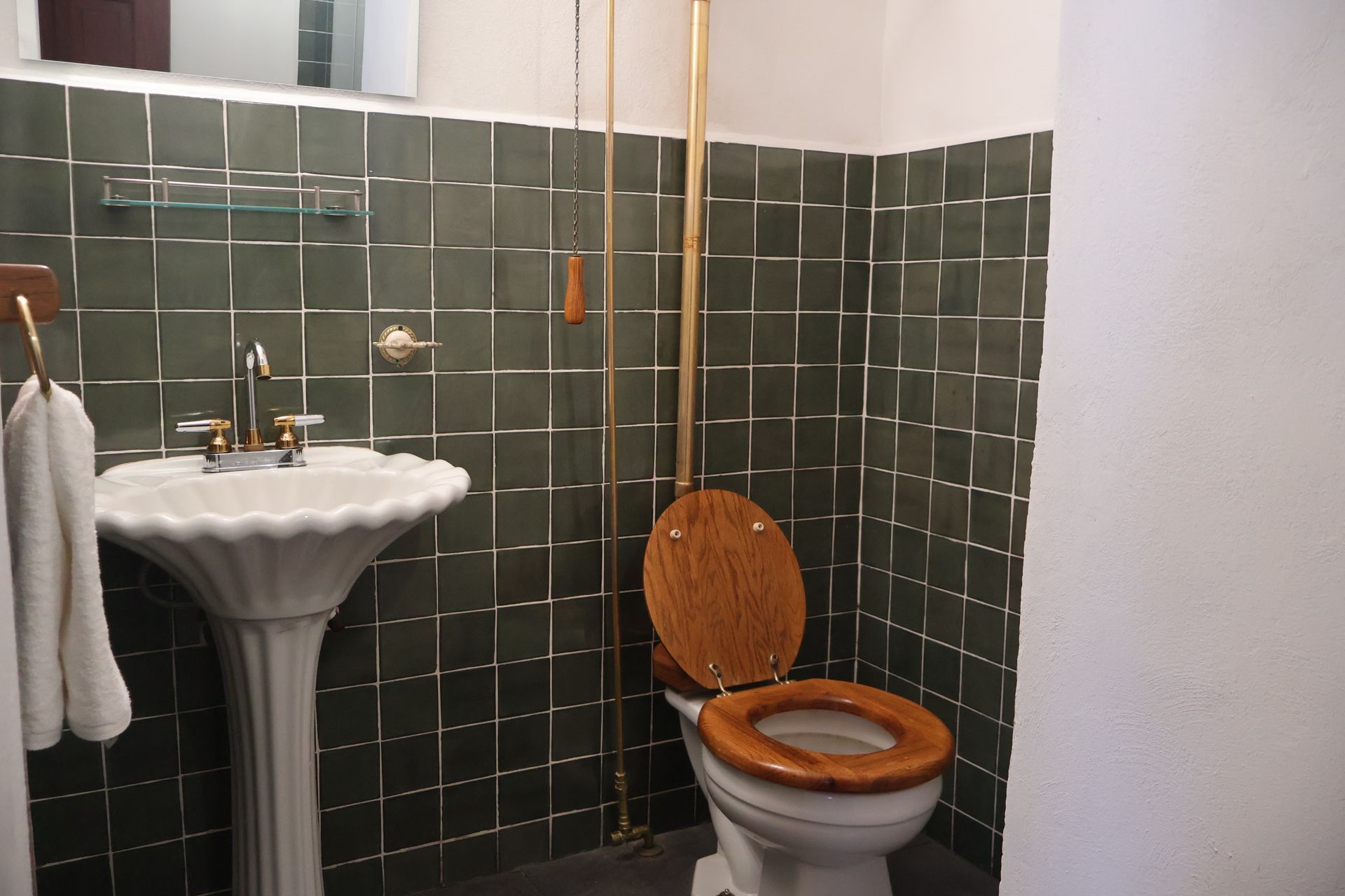 Baño con paredes de azulejos de color verde oscuro, lavabo con pedestal e inodoro con cadena.