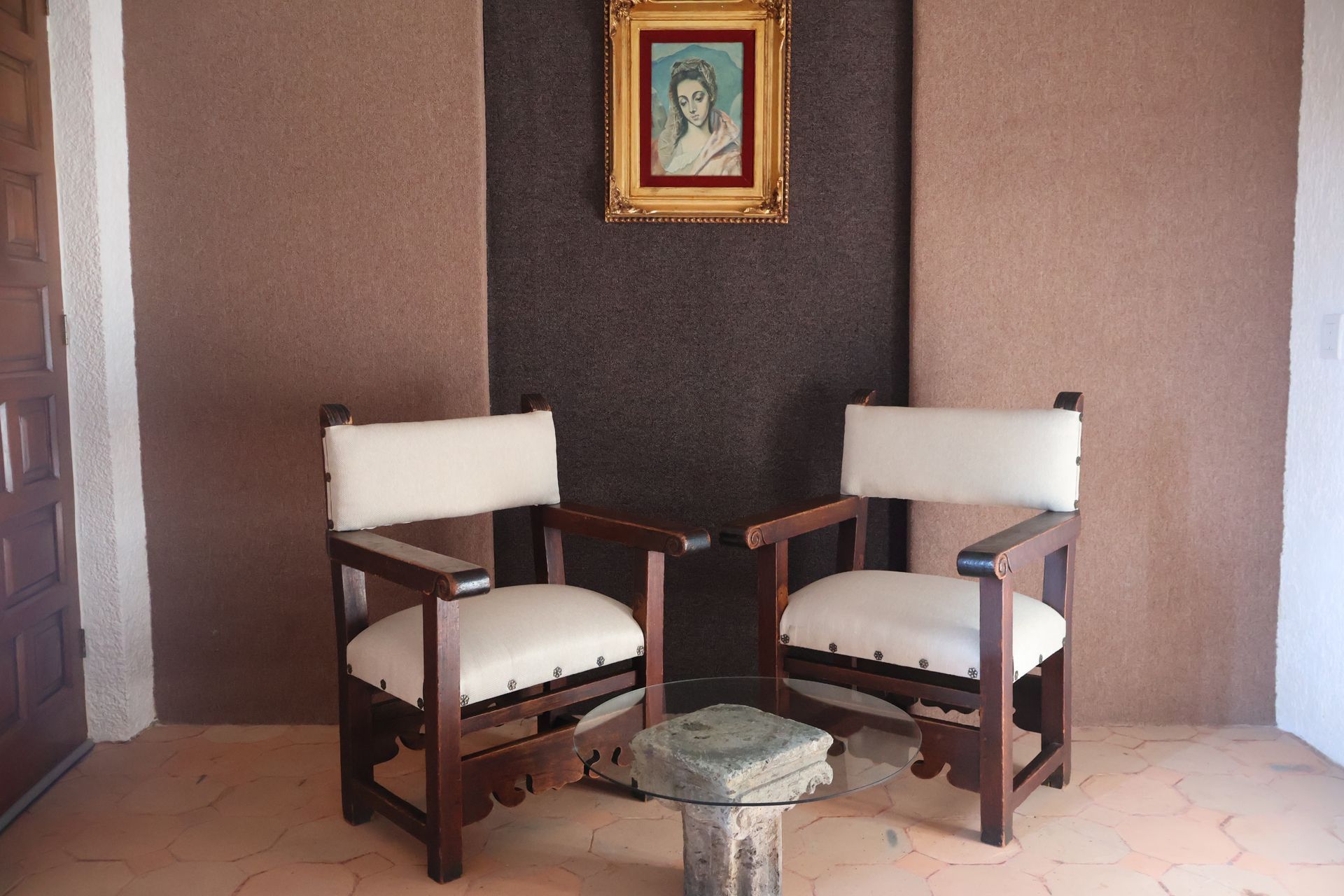 Dos sillas con asientos blancos, mesa pequeña, debajo de un cuadro sobre una pared marrón.